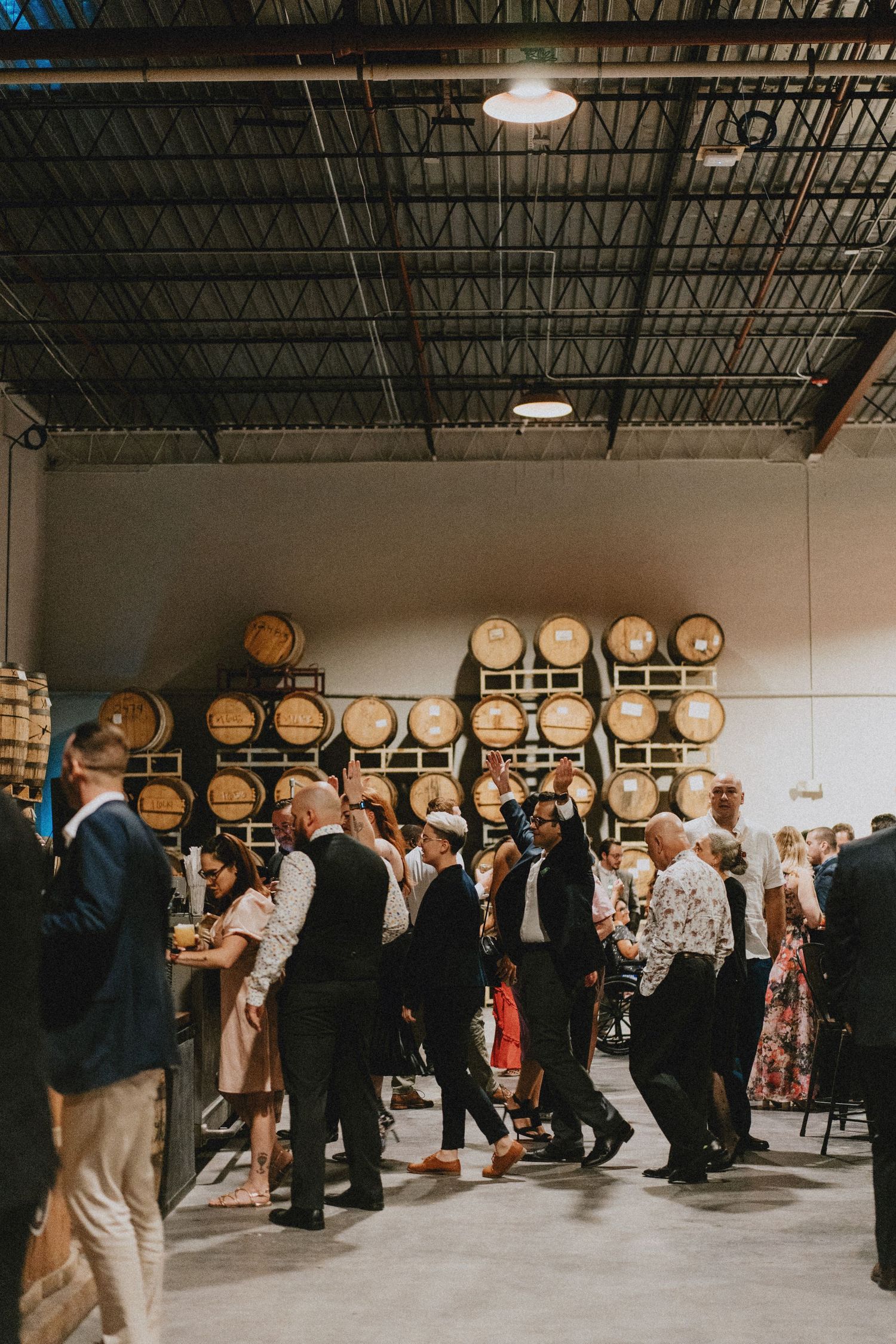 Guests mingle at a winery event venue with wooden barrels stacked on the wall in an industrial-style space.