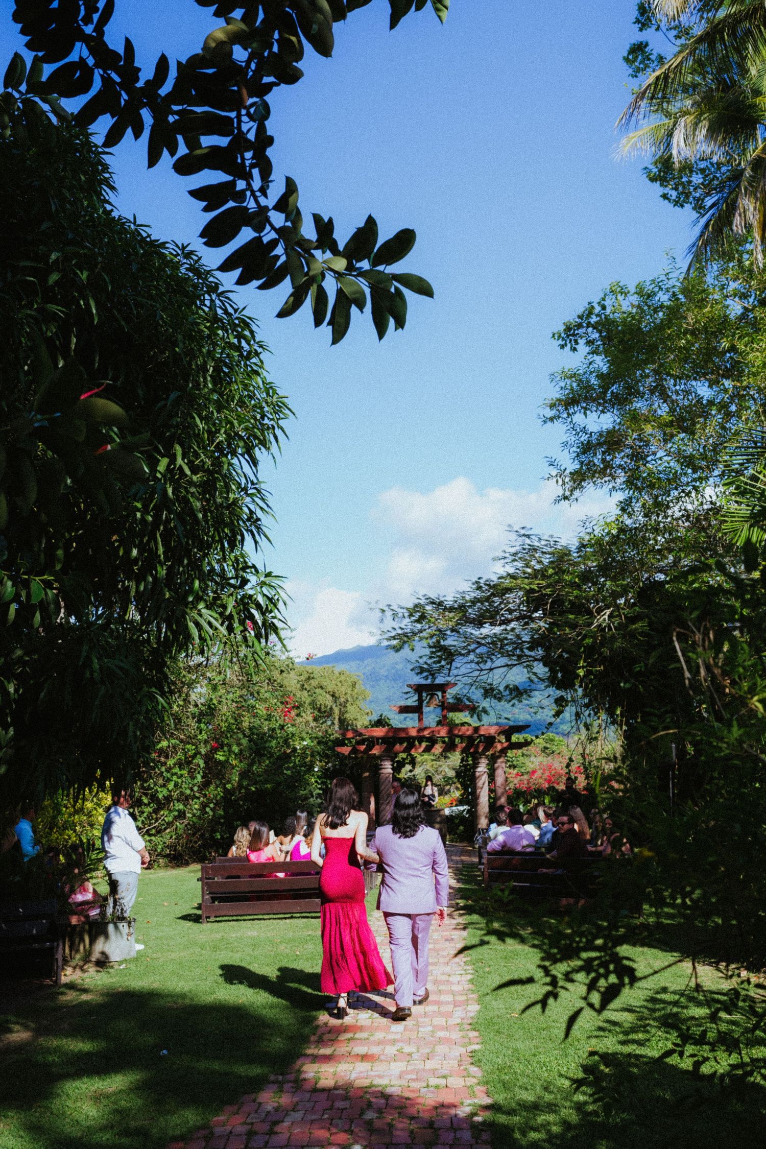 Couple walking hand in hand through a lush tropical garden with mountain views and guests gathered under a wooden pergola.