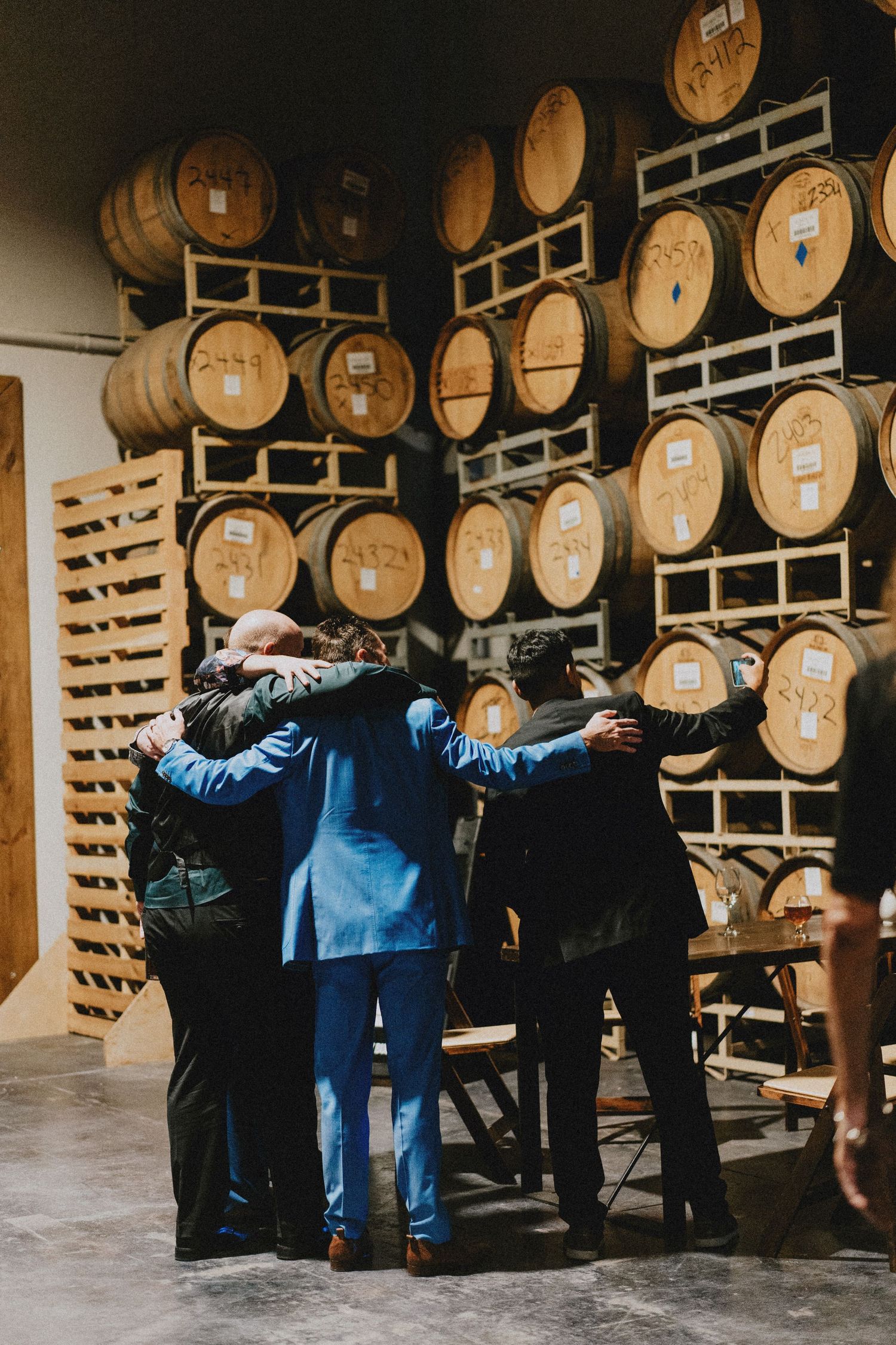 Groomsmen share a group hug in front of wooden wine barrels at a winery wedding venue.