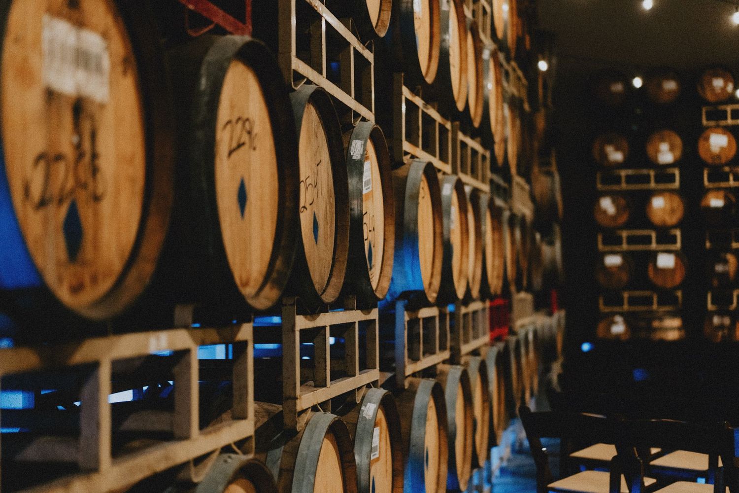 Rows of oak aging barrels stacked on wooden racks in a dimly lit distillery warehouse cellar.