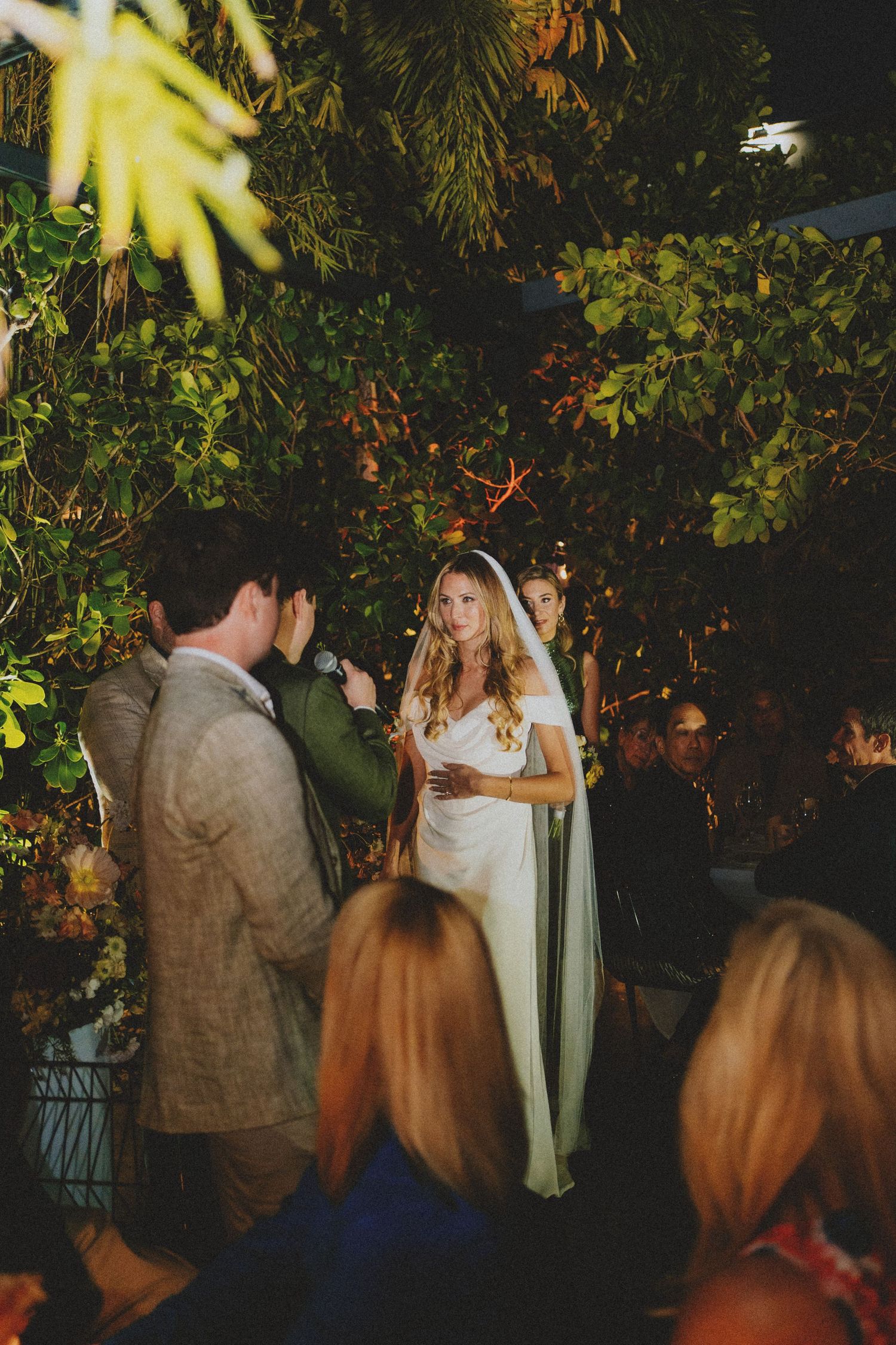 Bride in white gown and veil stands during outdoor wedding ceremony surrounded by lush greenery at night.