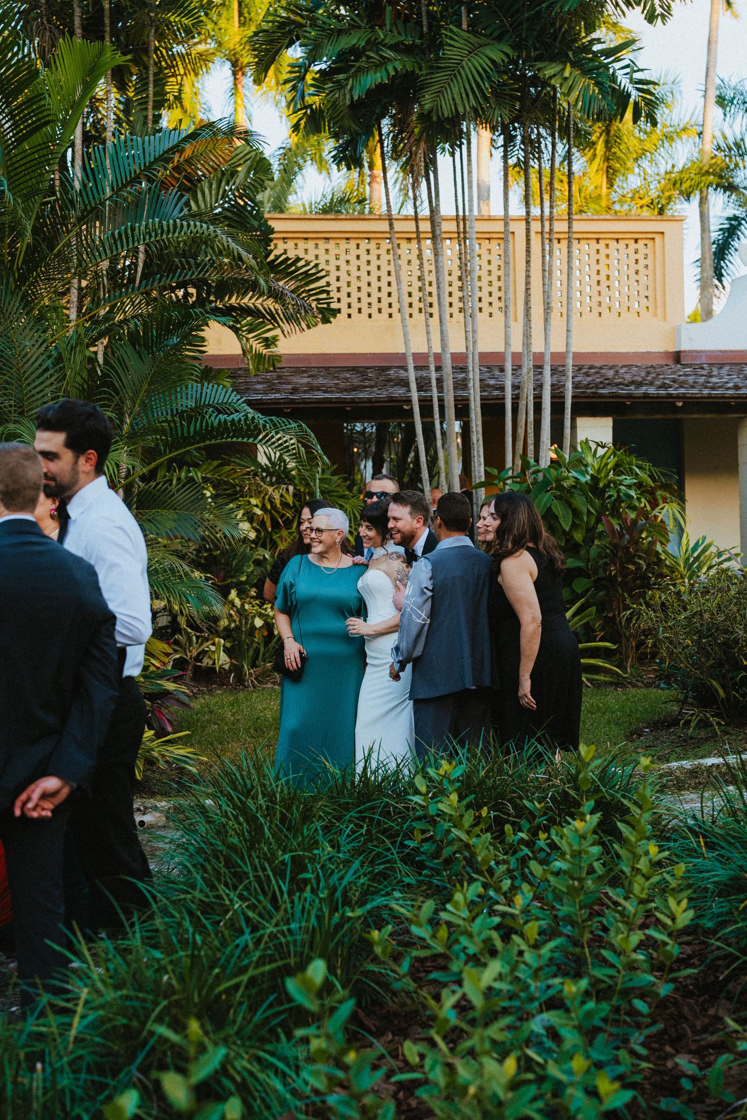 Wedding guests mingle outdoors among lush tropical greenery and tall palm trees at reception.
