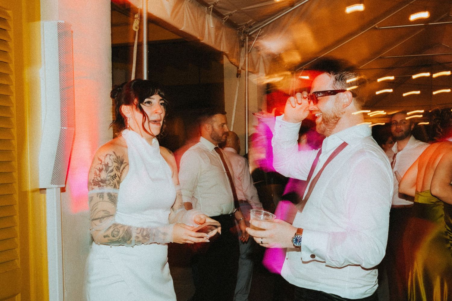 Tattooed bride in white dress dancing with guests at a lively wedding reception under a tent with pink lighting.