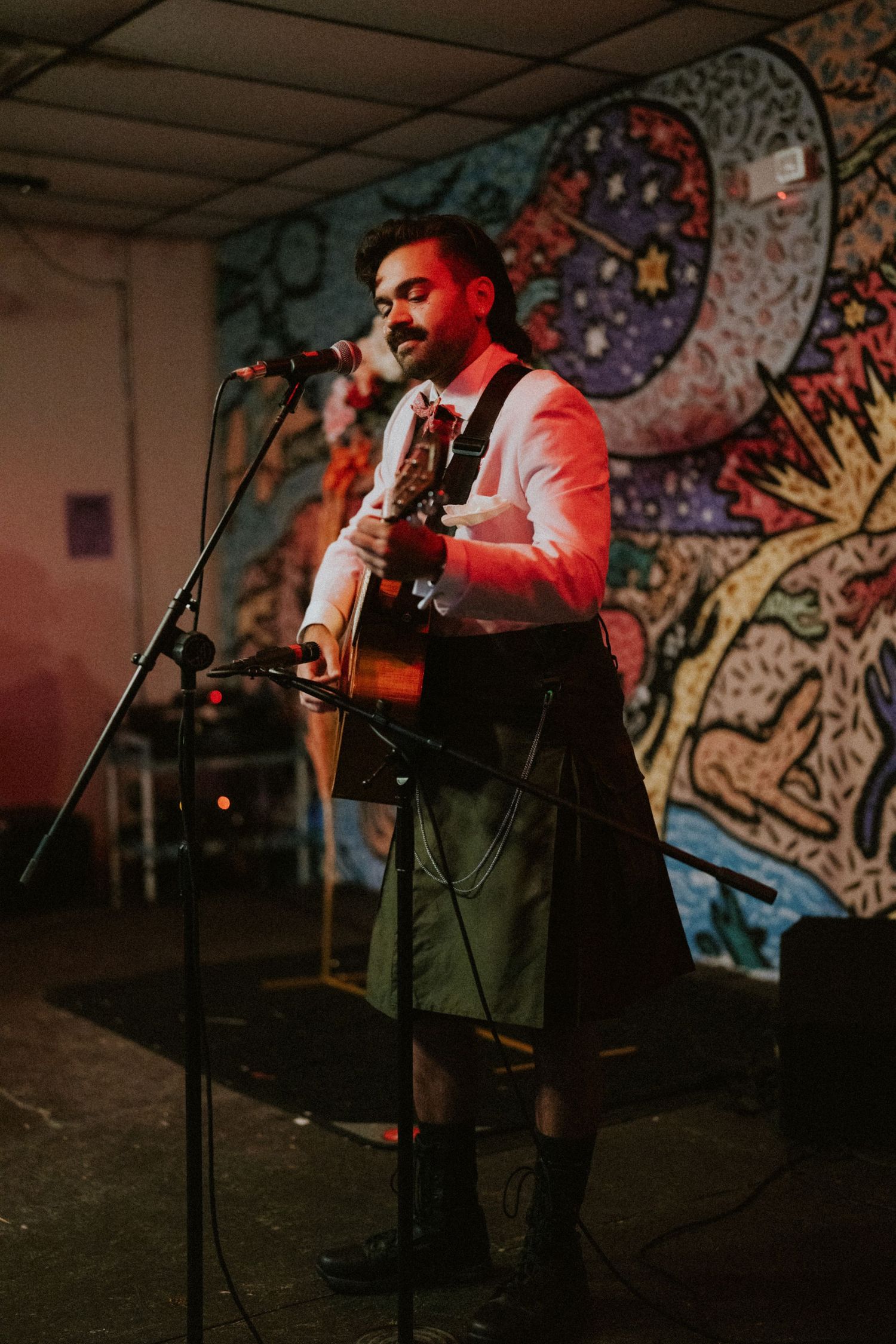 Bearded musician in kilt plays acoustic guitar at microphone stand before colorful mural backdrop.