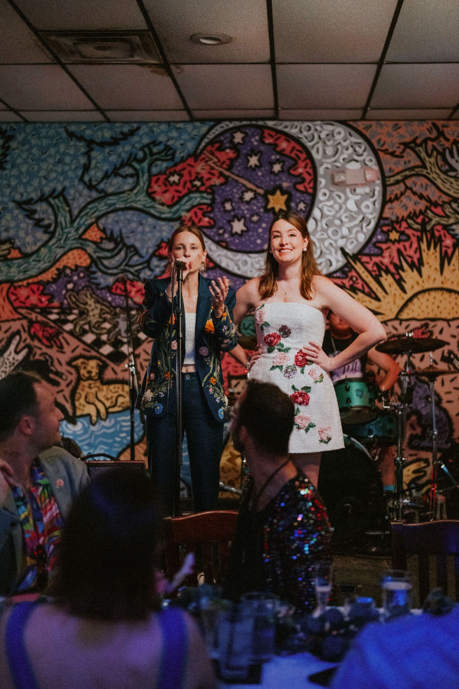 Two women stand on stage before vibrant psychedelic mural during wedding reception celebration.