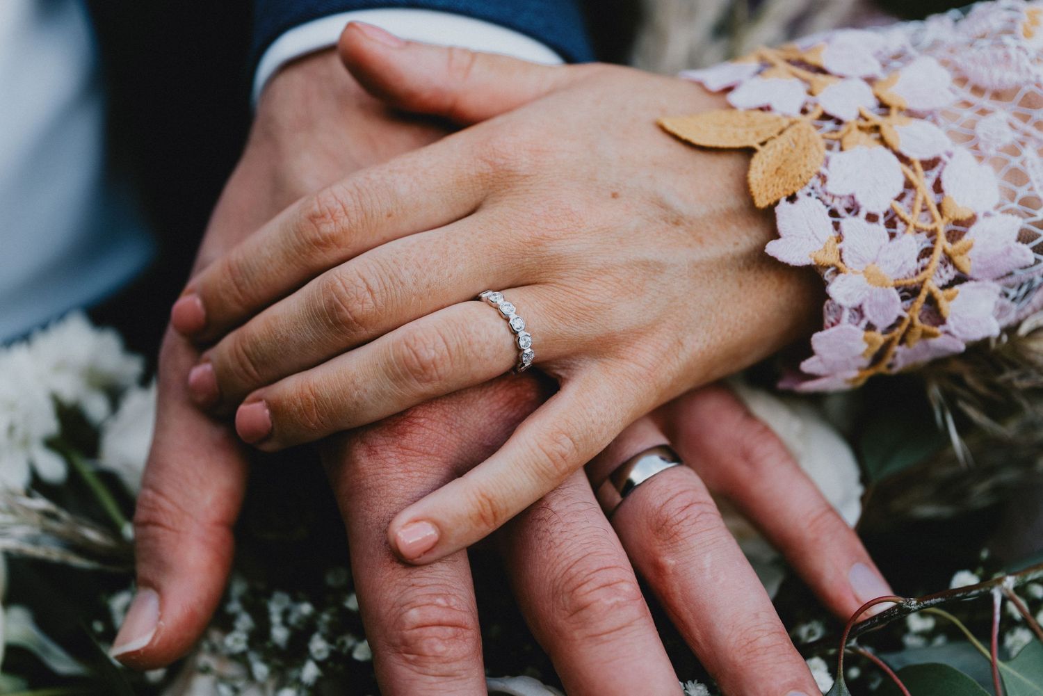 Newlywed couple displaying wedding rings on stacked hands over bridal bouquet flowers.