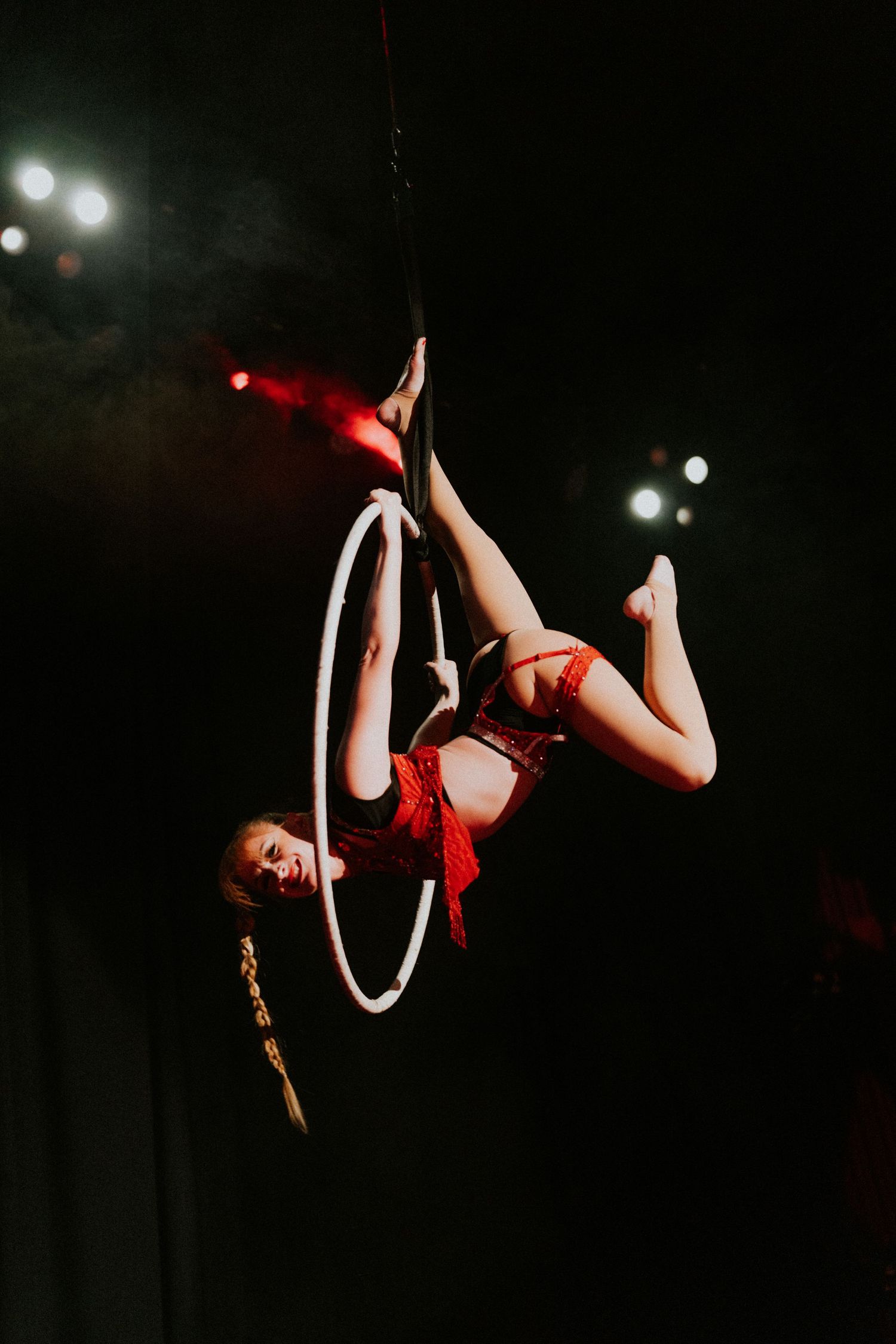 Aerial acrobat performing in a white hoop lyra against a dark stage backdrop in a red costume.