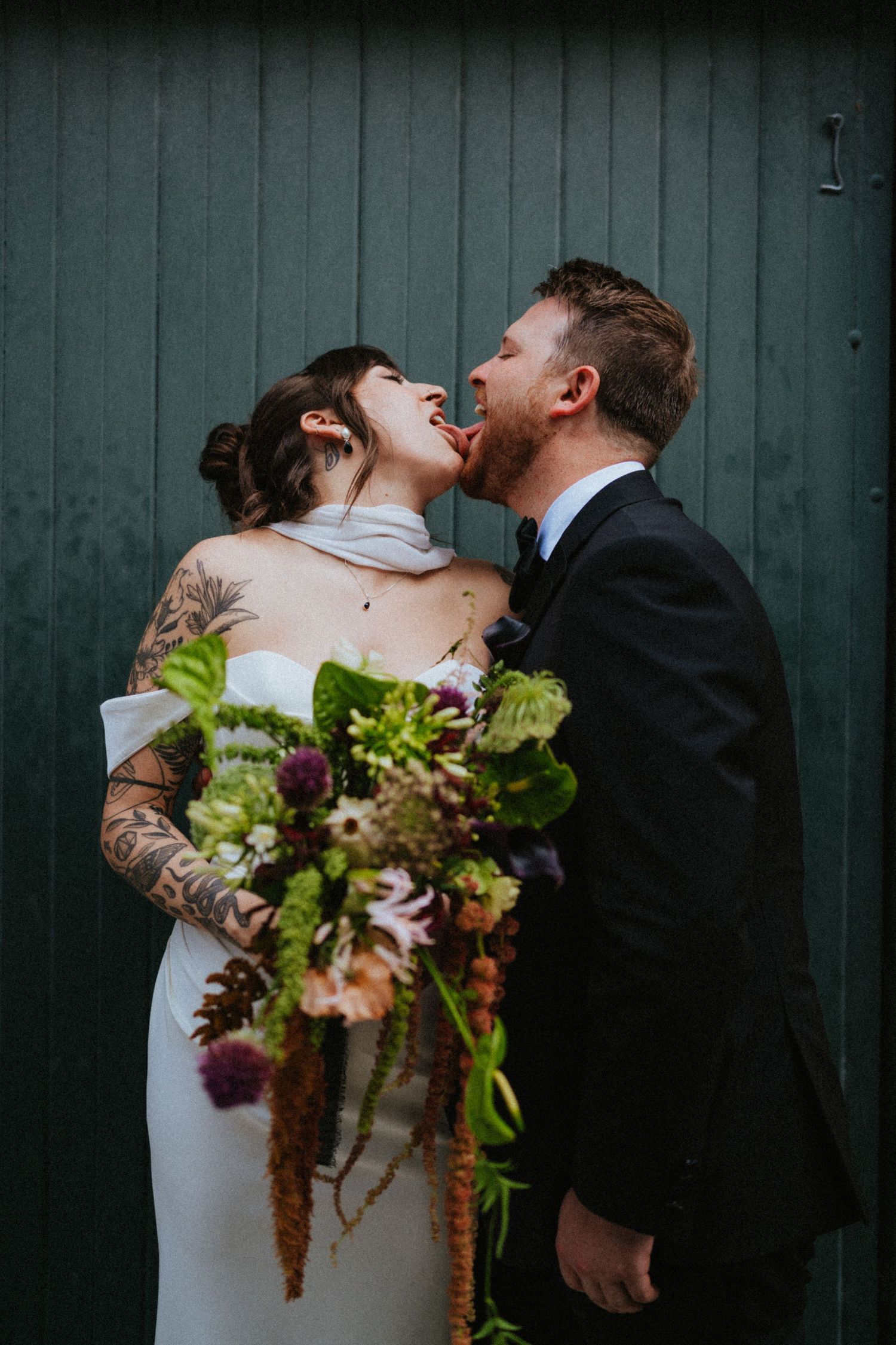 Bride with tattoos and groom in black suit laughing and kissing, holding lush green wedding bouquet against dark teal barn door.