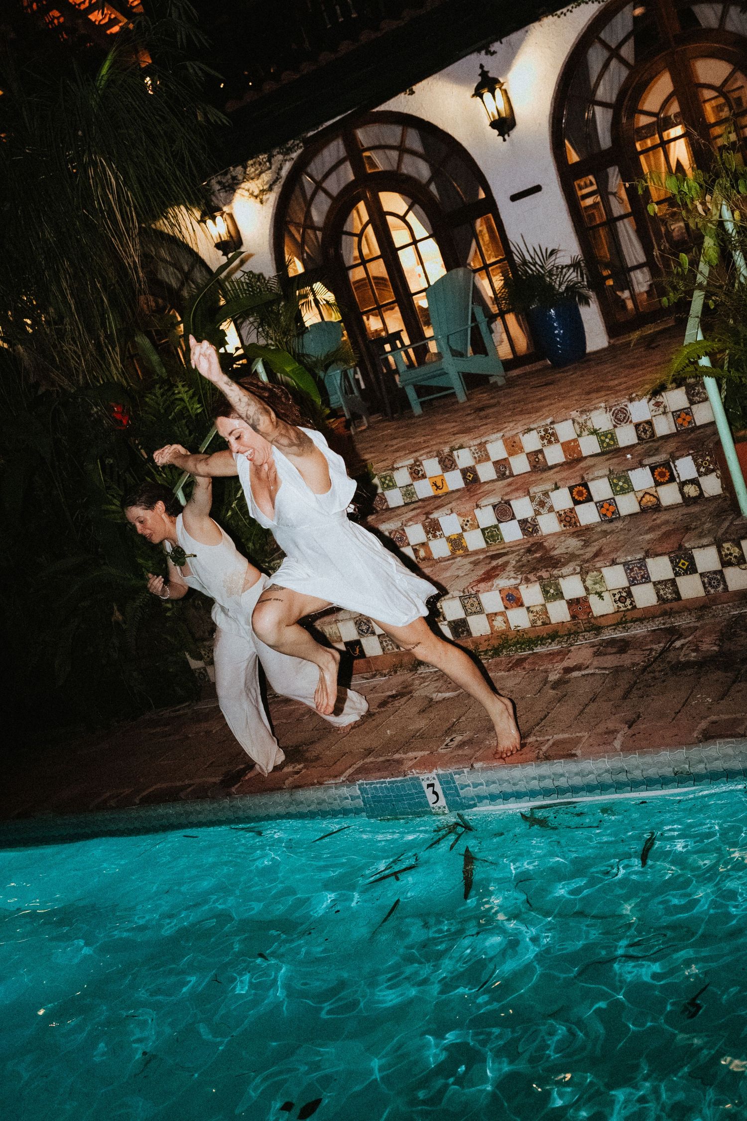 Bride and groom in white attire joyfully jumping into a pool at night during wedding celebration.