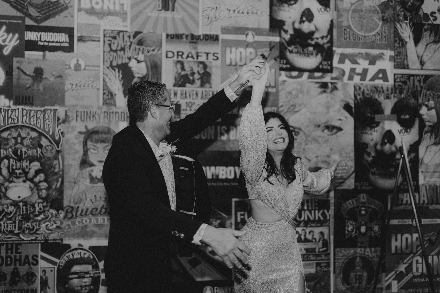 Couple dancing joyfully in black and white photo against a colorful vintage poster wall at their wedding reception.