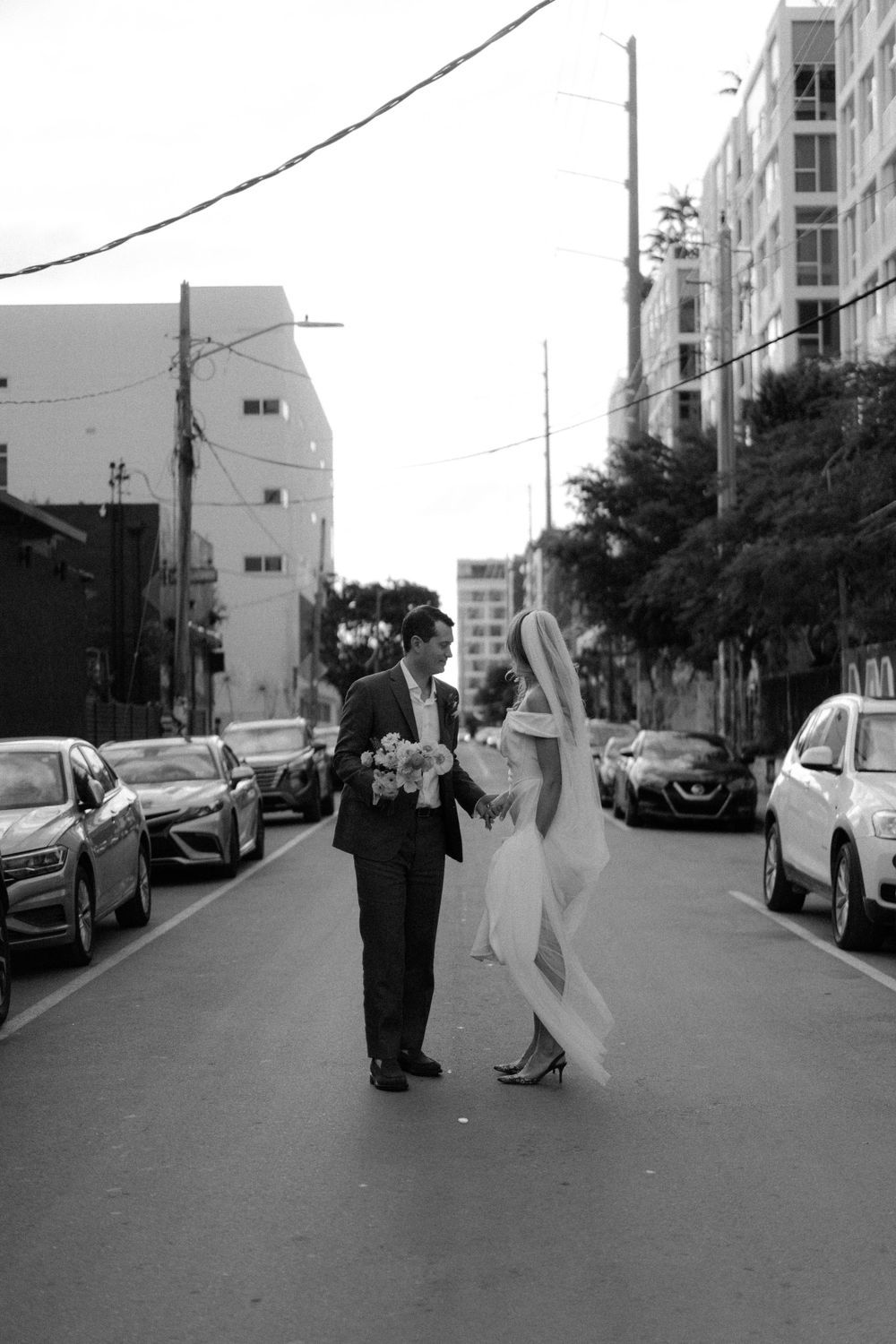 Black and white photo of newlyweds dancing in a city street lined with parked cars.