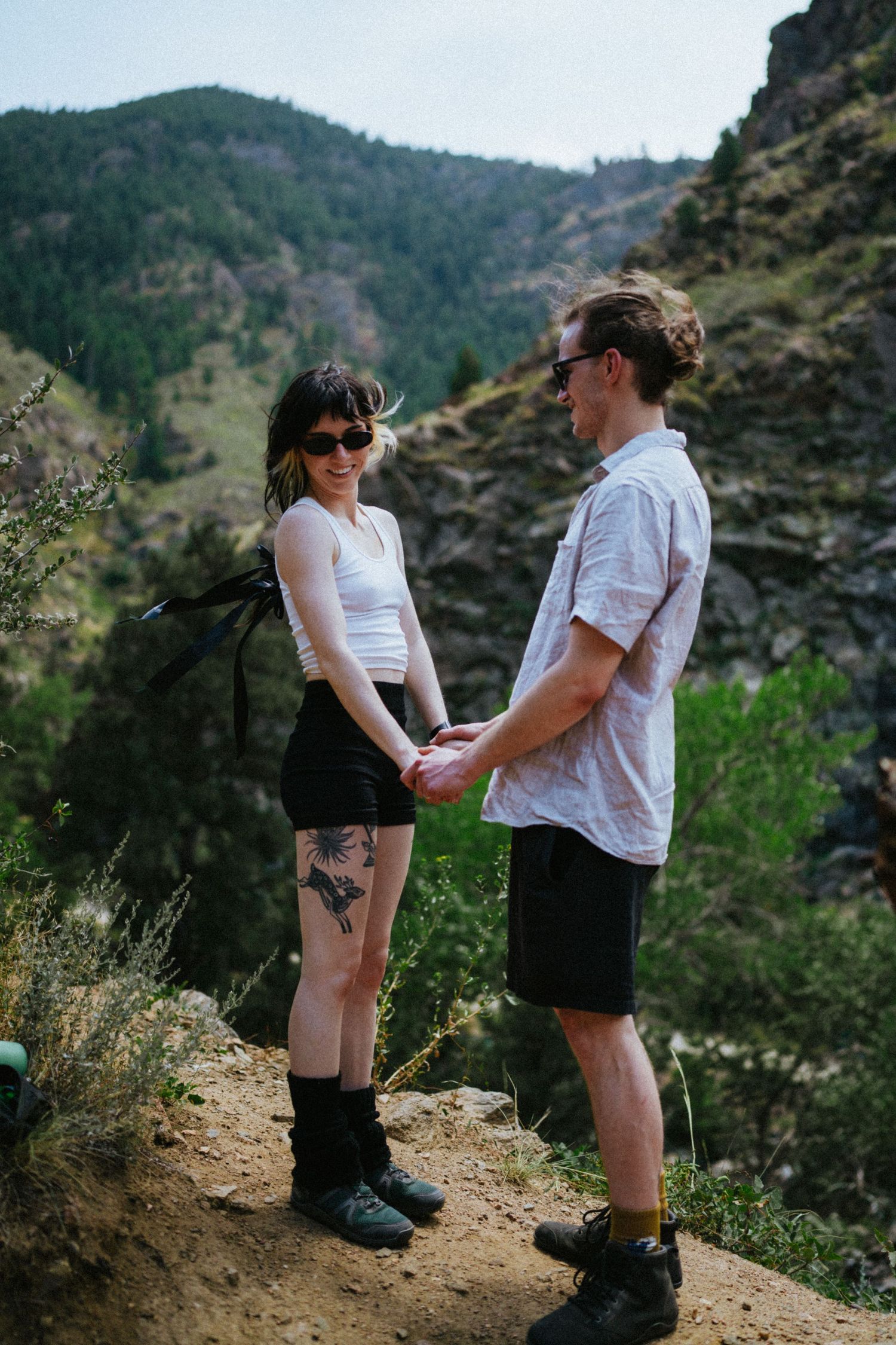 Couple holding hands on a mountain hiking trail, woman with tattoos in white tank top and man in white shirt, scenic rocky landscape.