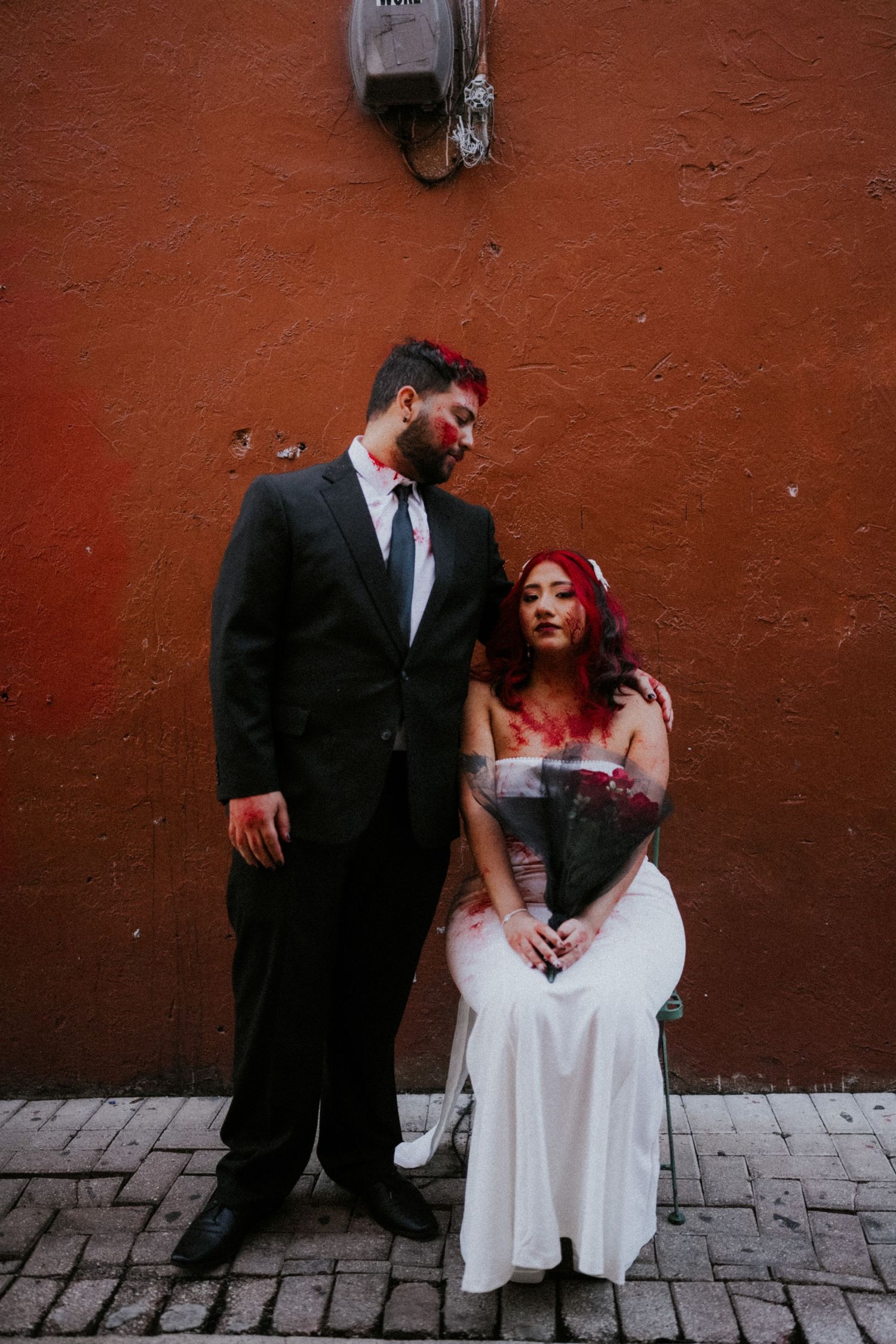 Zombie wedding couple with fake blood posing against rustic red wall, groom in black suit, bride seated in white gown.