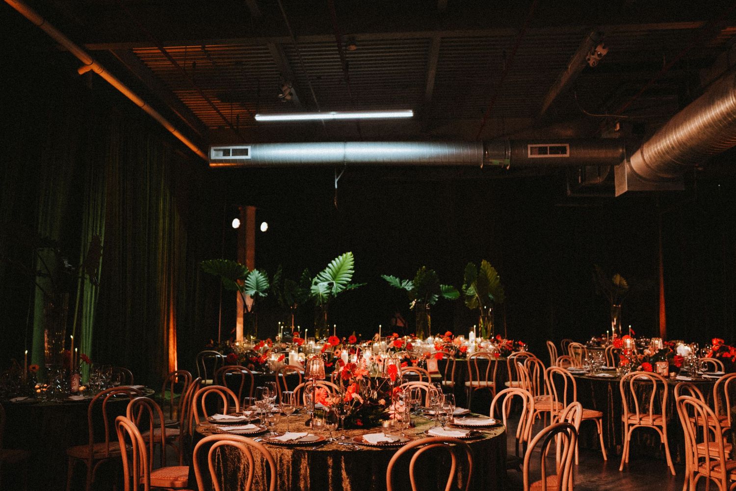 Elegant wedding reception in dark industrial venue with round tables, bentwood chairs, red floral centerpieces, and tropical plant backdrop.