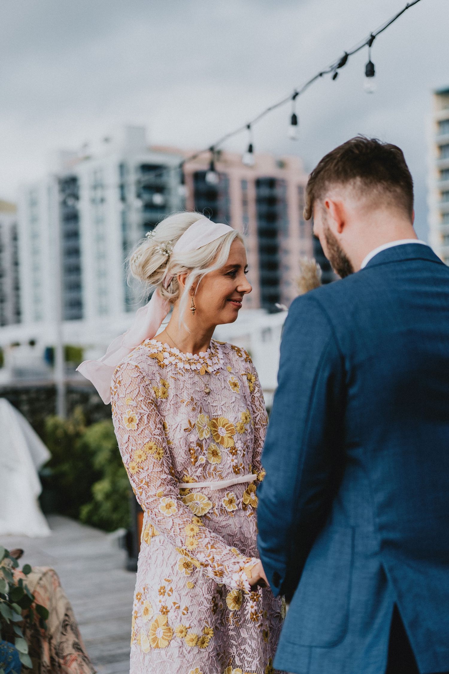 Bride in floral embroidered gown exchanges vows with groom in blue suit on rooftop venue with city skyline backdrop.