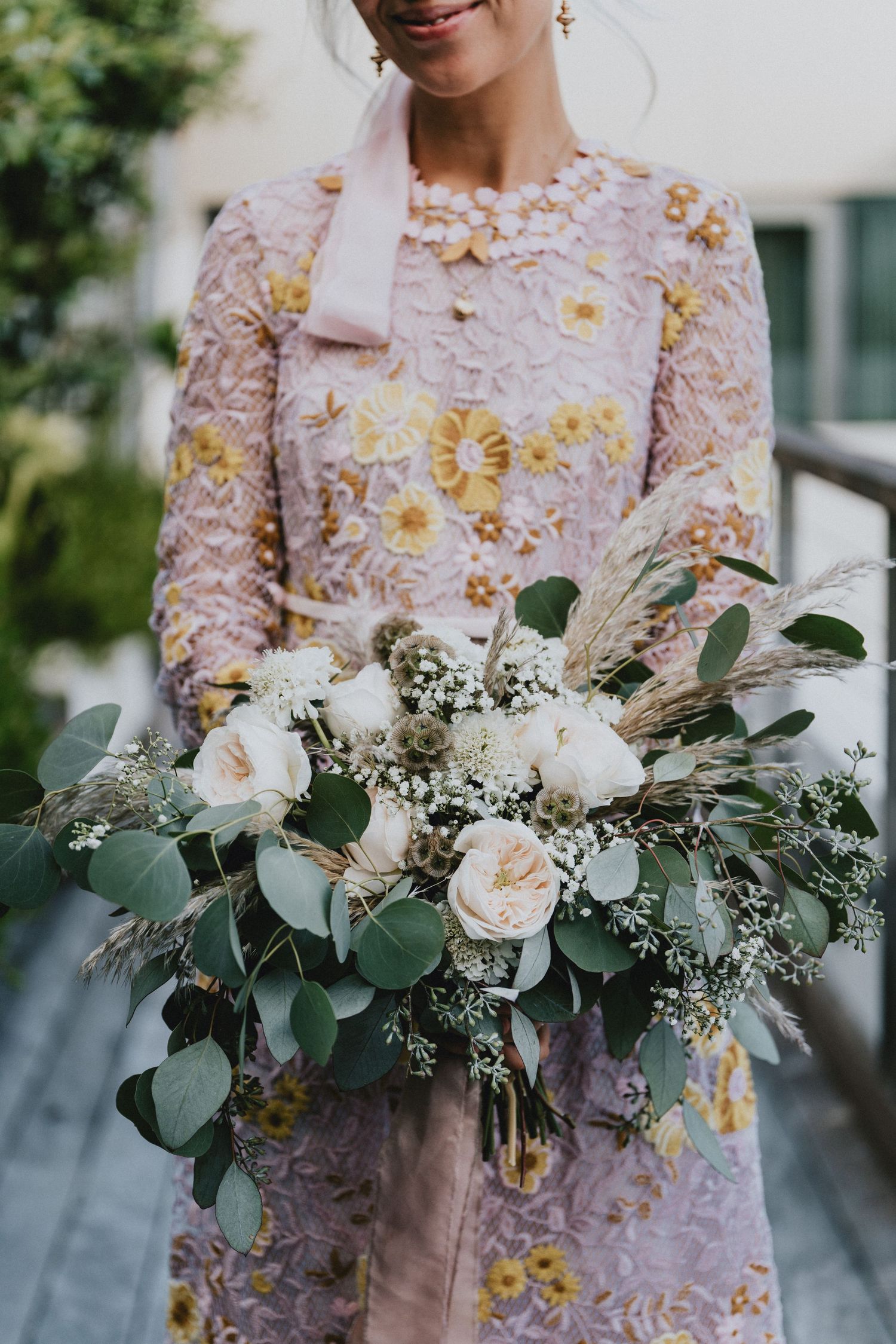 Bride in pink floral lace dress holding a lush bouquet of white roses, eucalyptus, and pampas grass.