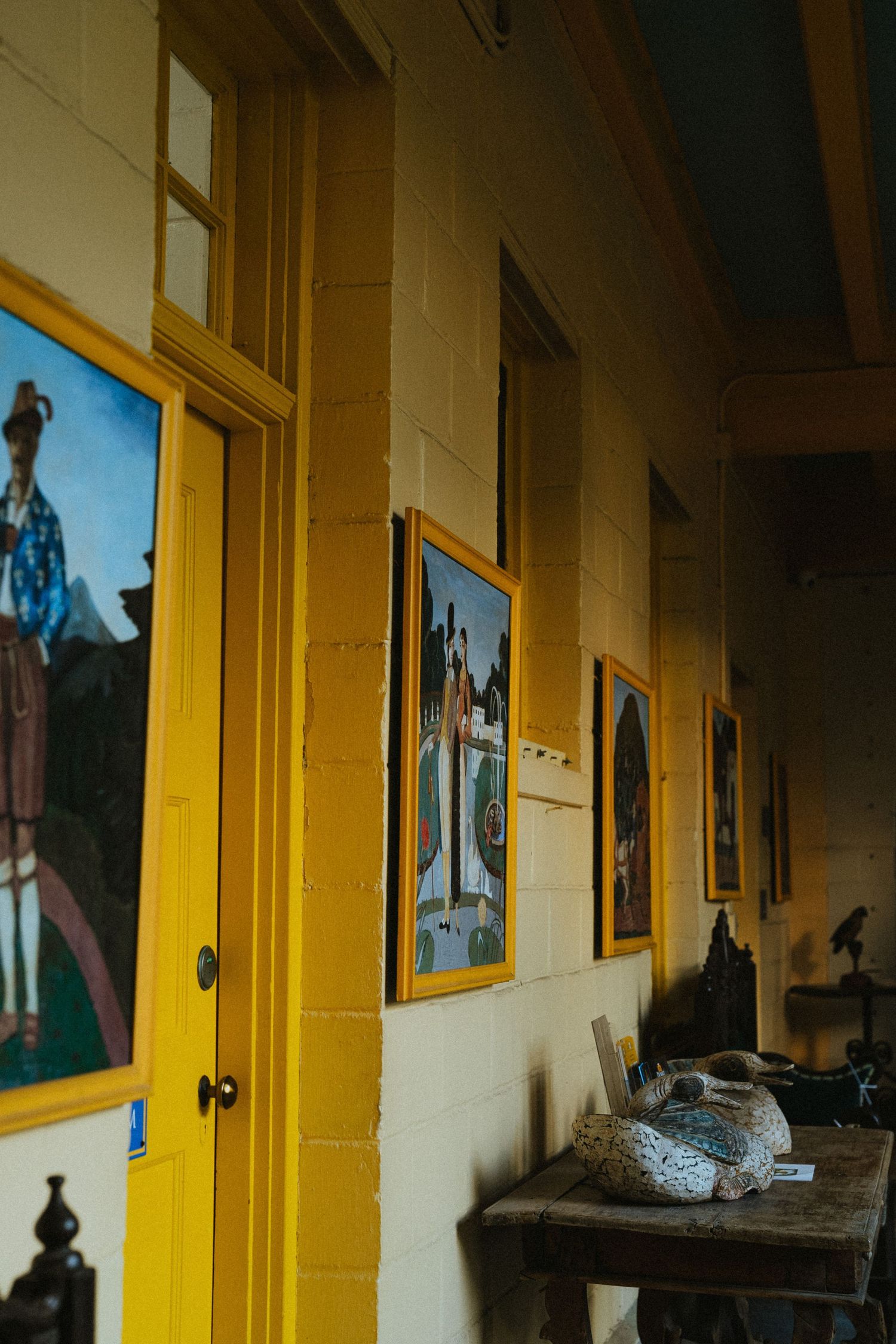 Yellow-framed paintings line a rustic gallery hallway with stone walls, bright doors, and a wooden table displaying decorative objects.