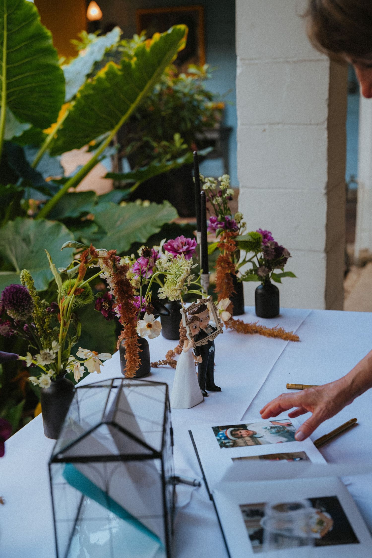 Wedding reception table decorated with wildflowers, cake topper figurines, and photo guest book display.