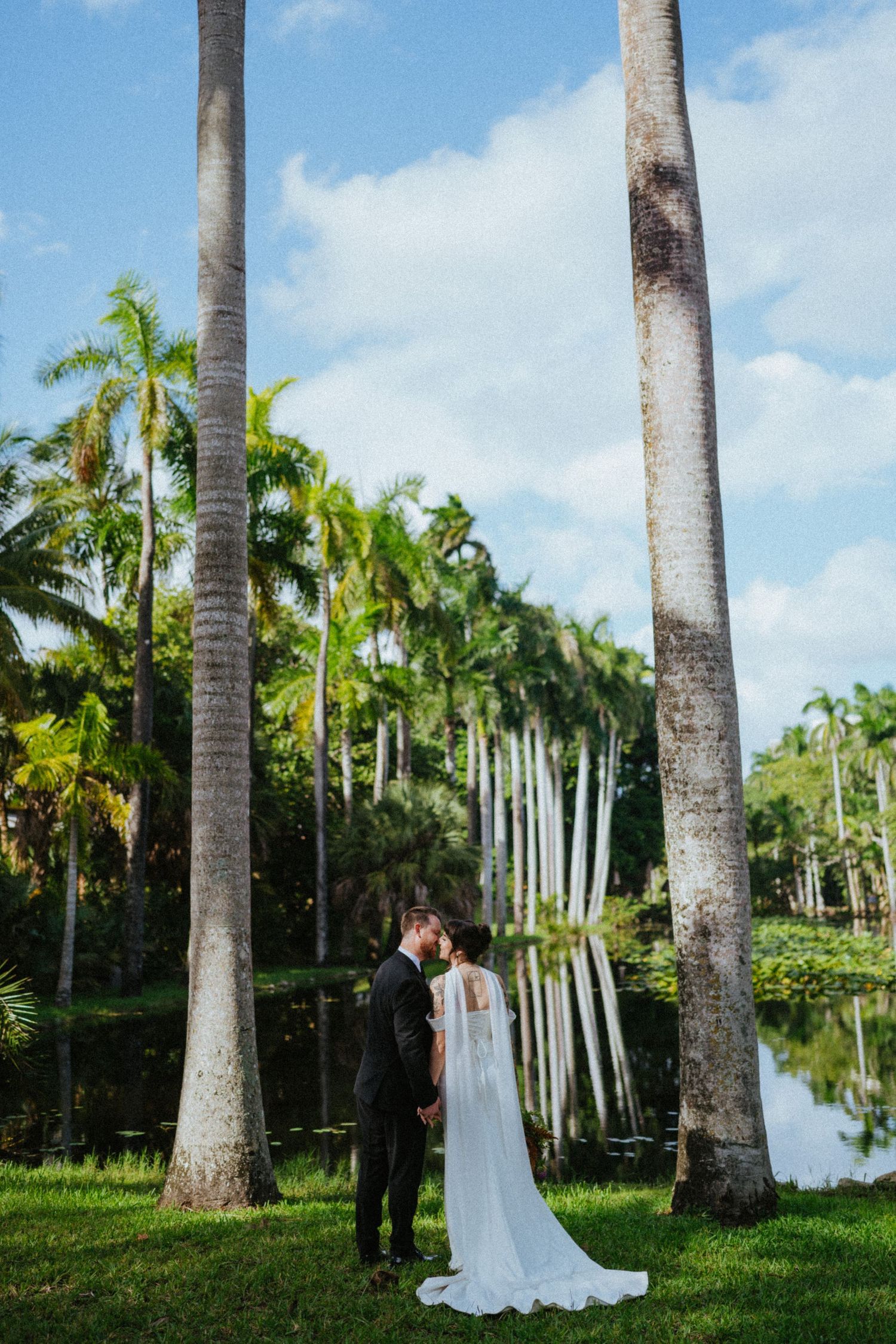 Bride and groom share a romantic kiss among tall palm trees beside a tranquil pond at a tropical outdoor wedding.