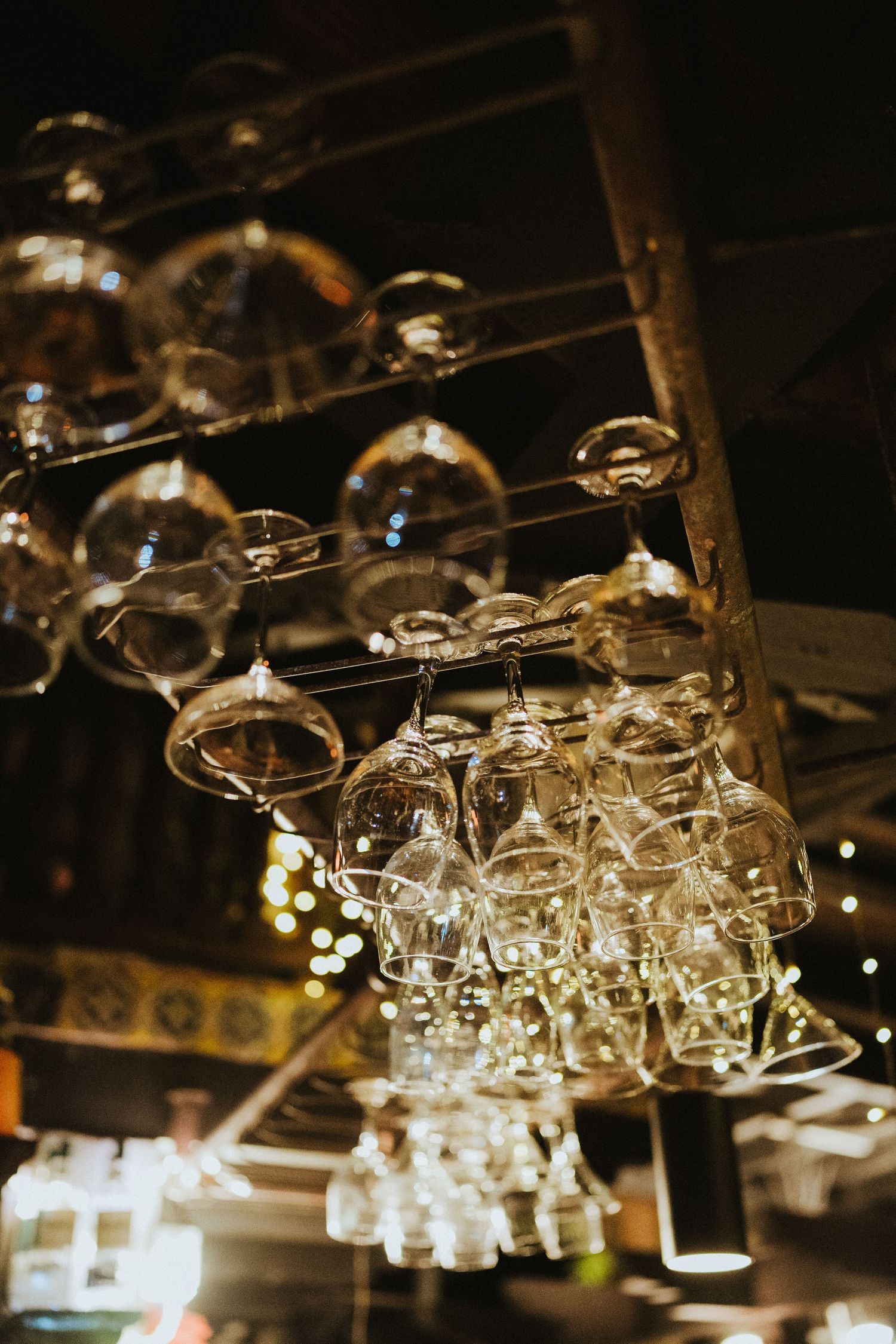 Wine glasses hanging upside down on a wooden rack above a dimly lit bar with warm bokeh lights in the background.