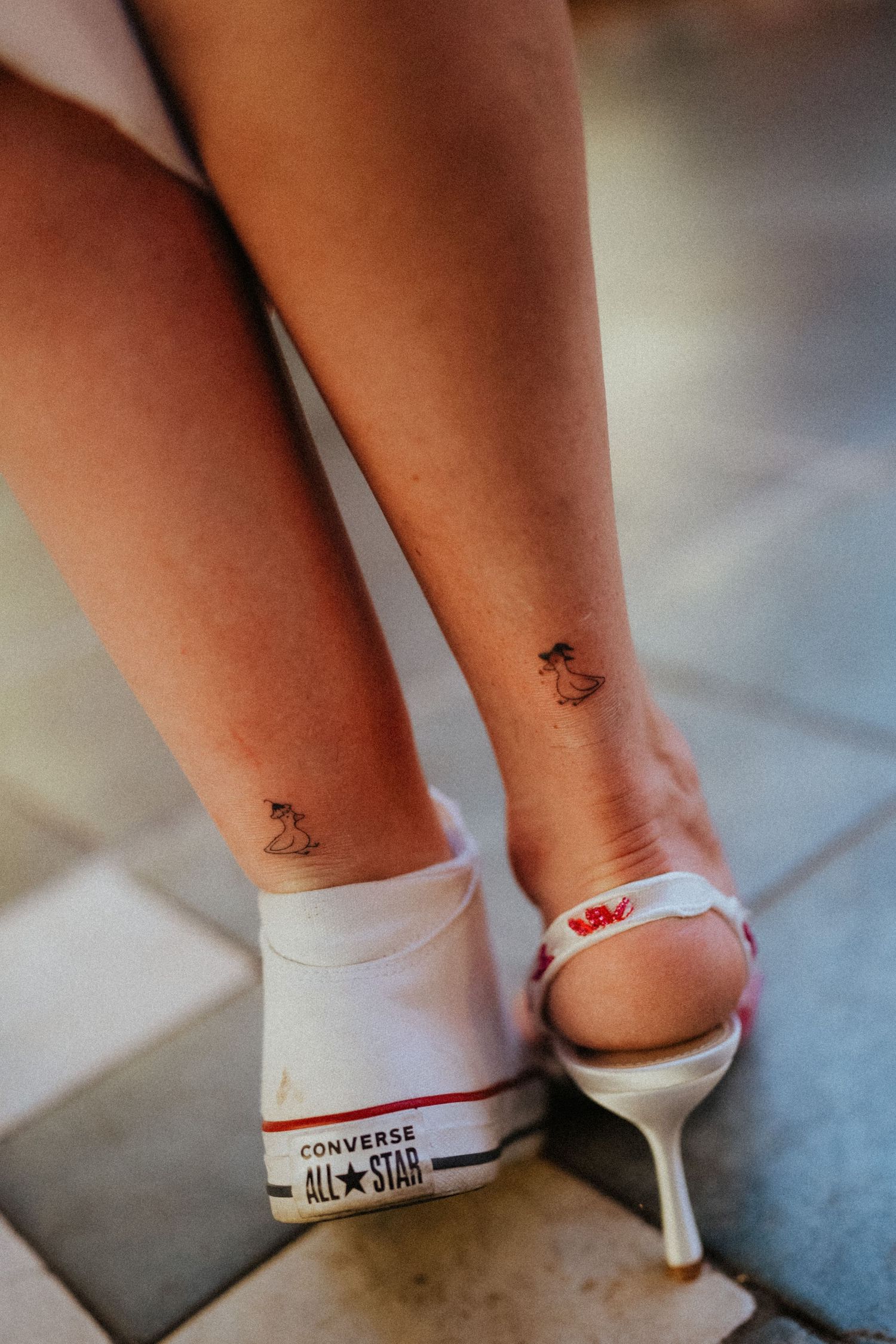 Close-up of ankles with small tattoos, one wearing a white Converse All Star sneaker and the other a white strappy heel.