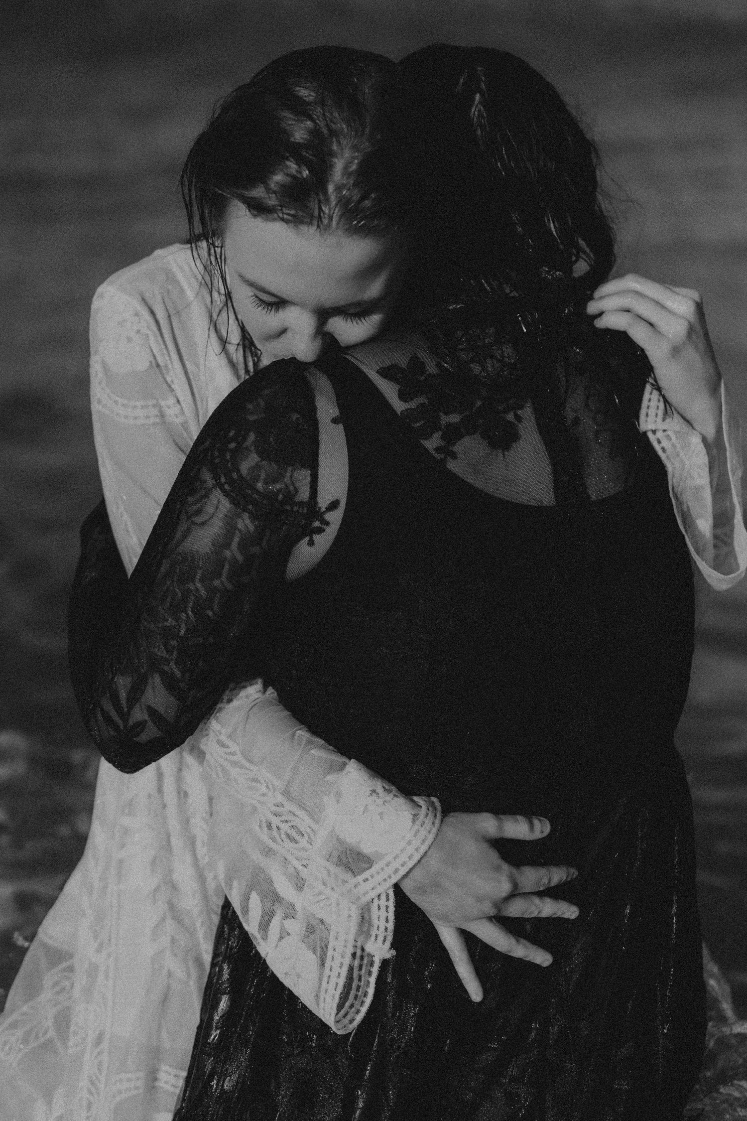 Black and white photo of two women in lace dresses embracing tenderly near water.