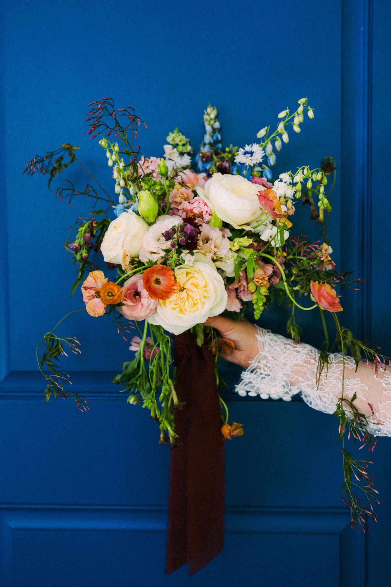 Hand holding a lush bridal bouquet with white, peach, and pink blooms, greenery, and a dark ribbon against a blue door.