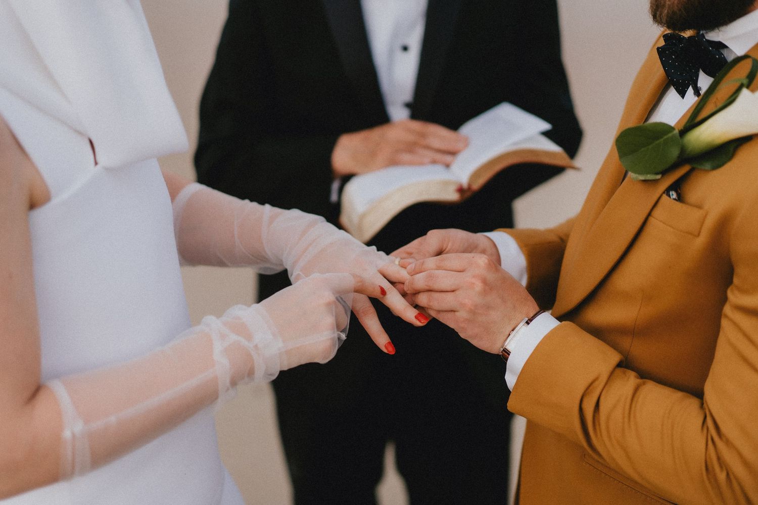 Bride placing wedding ring on groom's finger during ceremony, officiant holding book in background.