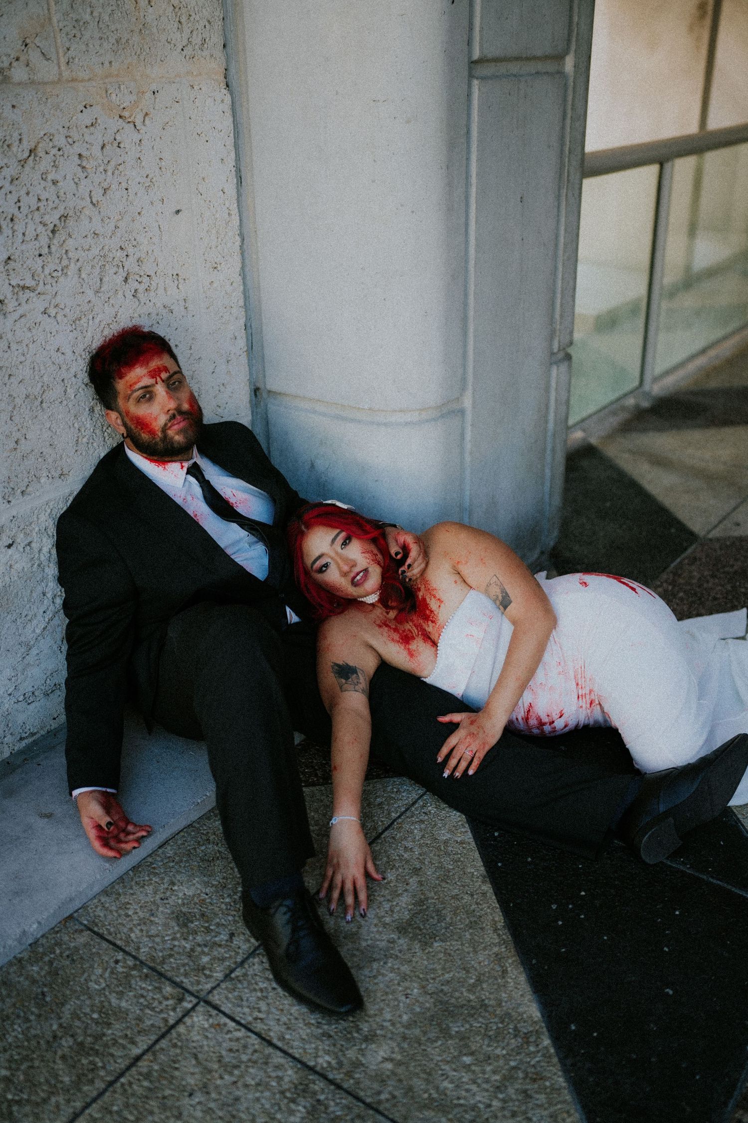 Couple in zombie Halloween costumes with fake blood, wearing wedding attire, posing dramatically on ground outside building.