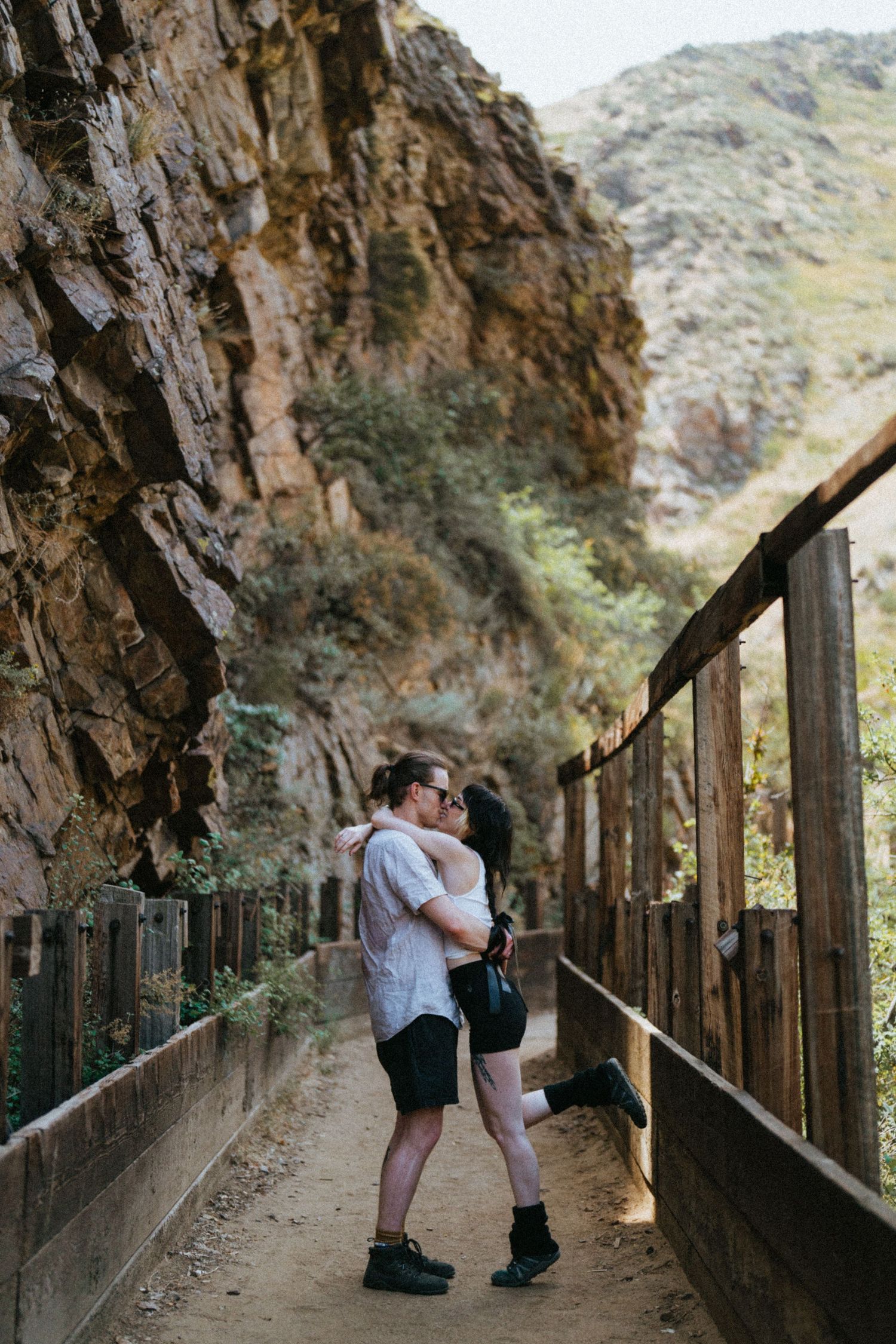 Couple sharing a romantic kiss on a wooden walkway bridge with rocky canyon walls and green hills backdrop.