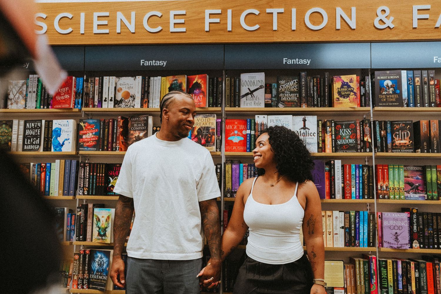 A smiling couple holds hands in front of a Science Fiction and Fantasy bookstore section.