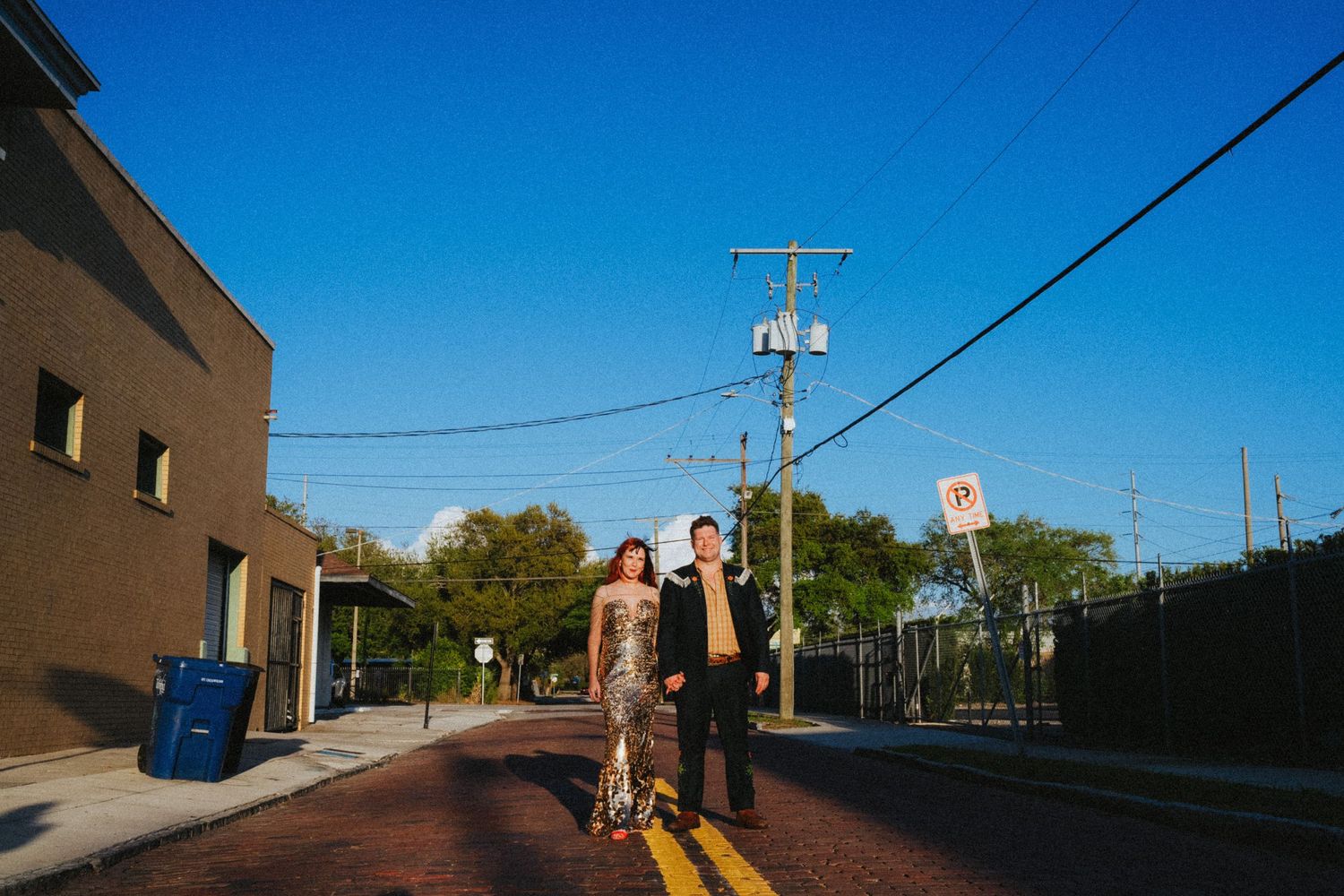 Couple standing on a brick street intersection under a clear blue sky with power lines and urban buildings in the background.