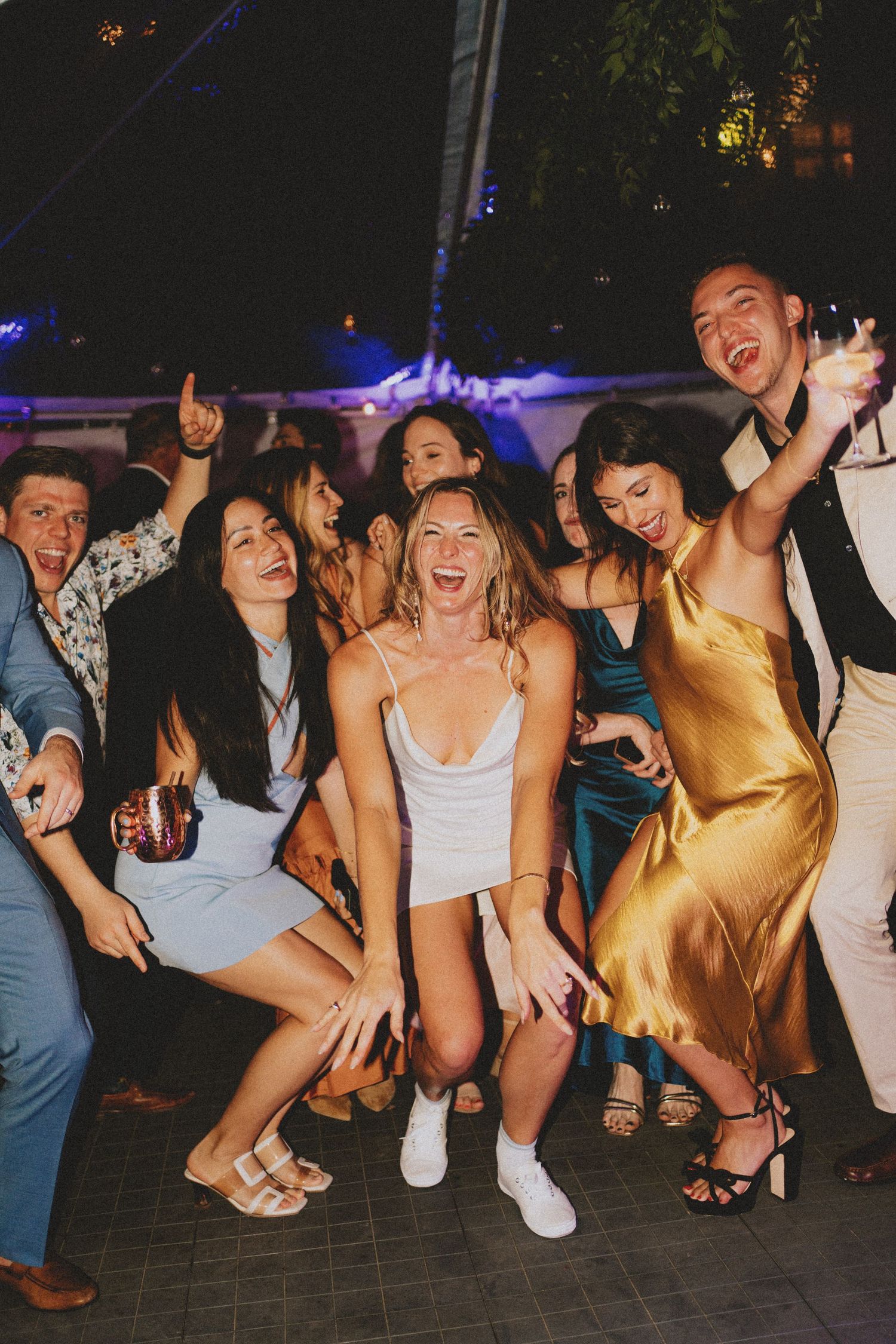 Group of joyful wedding guests dancing and posing together at an outdoor nighttime reception.