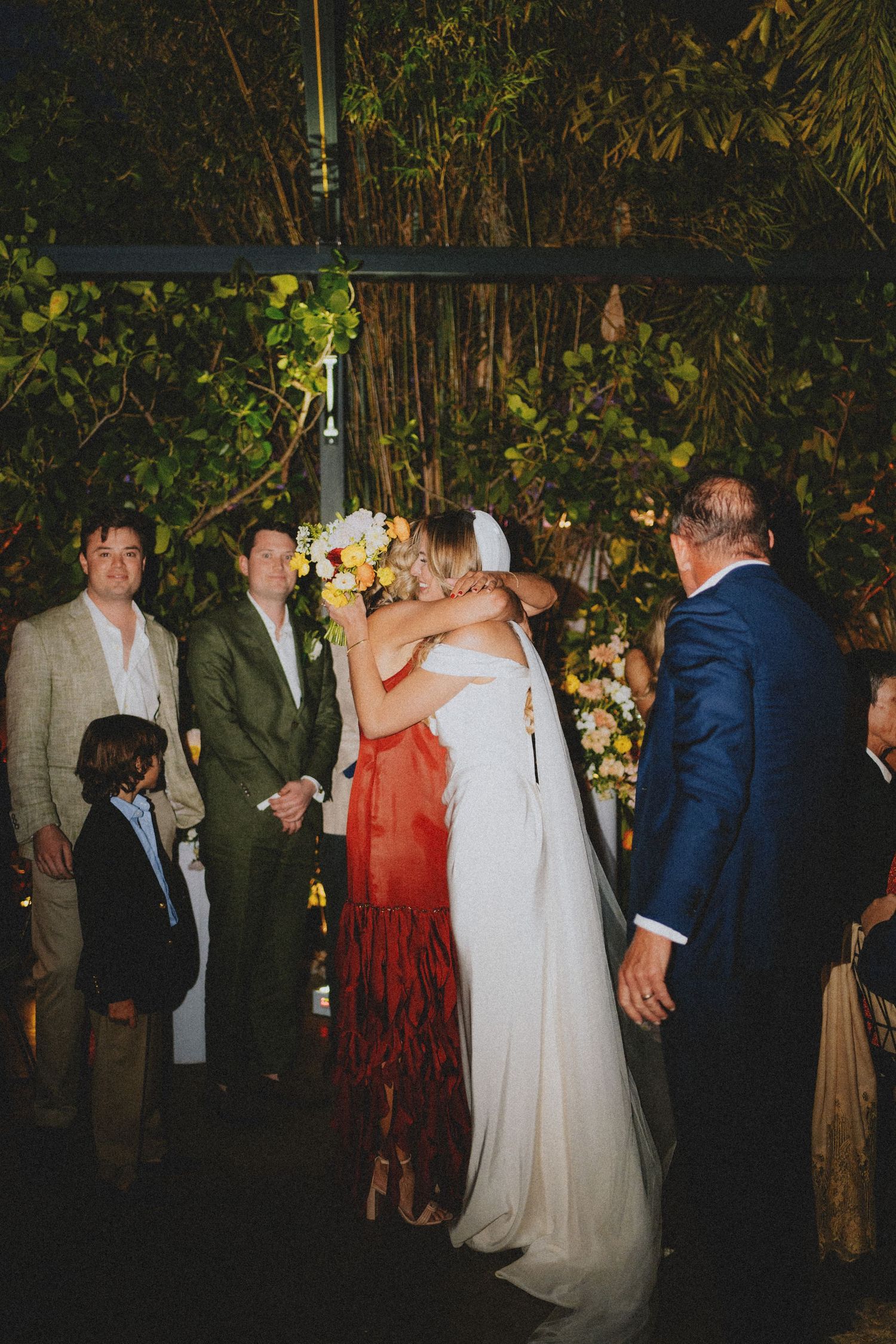 Bride embraces a guest during outdoor evening wedding ceremony surrounded by lush tropical greenery.
