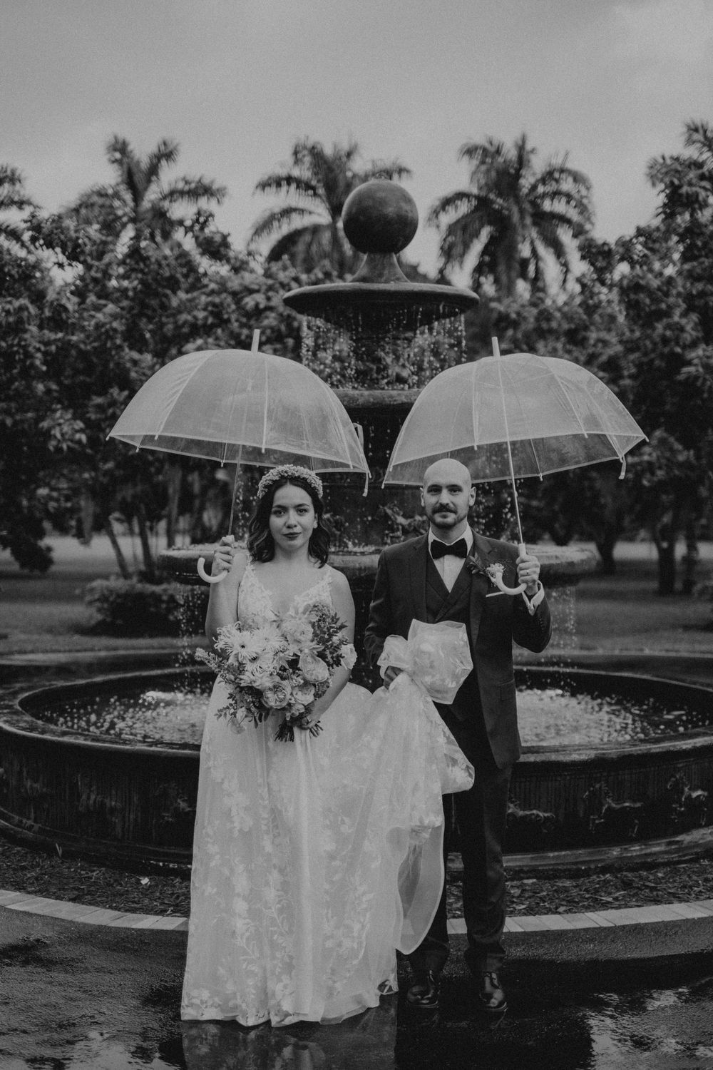 Couple poses with clear umbrellas in a rainy garden setting during their wedding photoshoot.