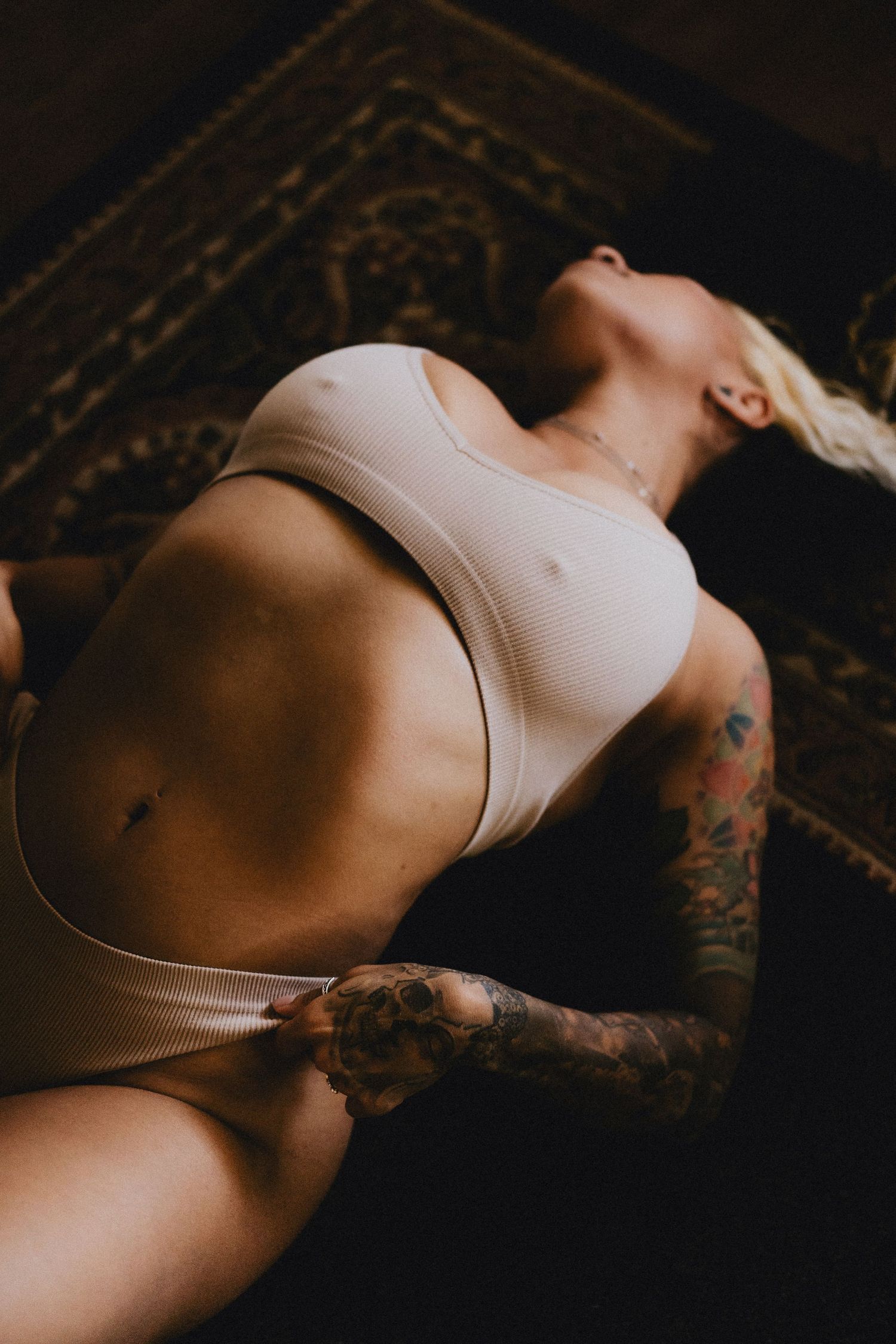 Person in beige lingerie with visible tattoos reclines on patterned rug.