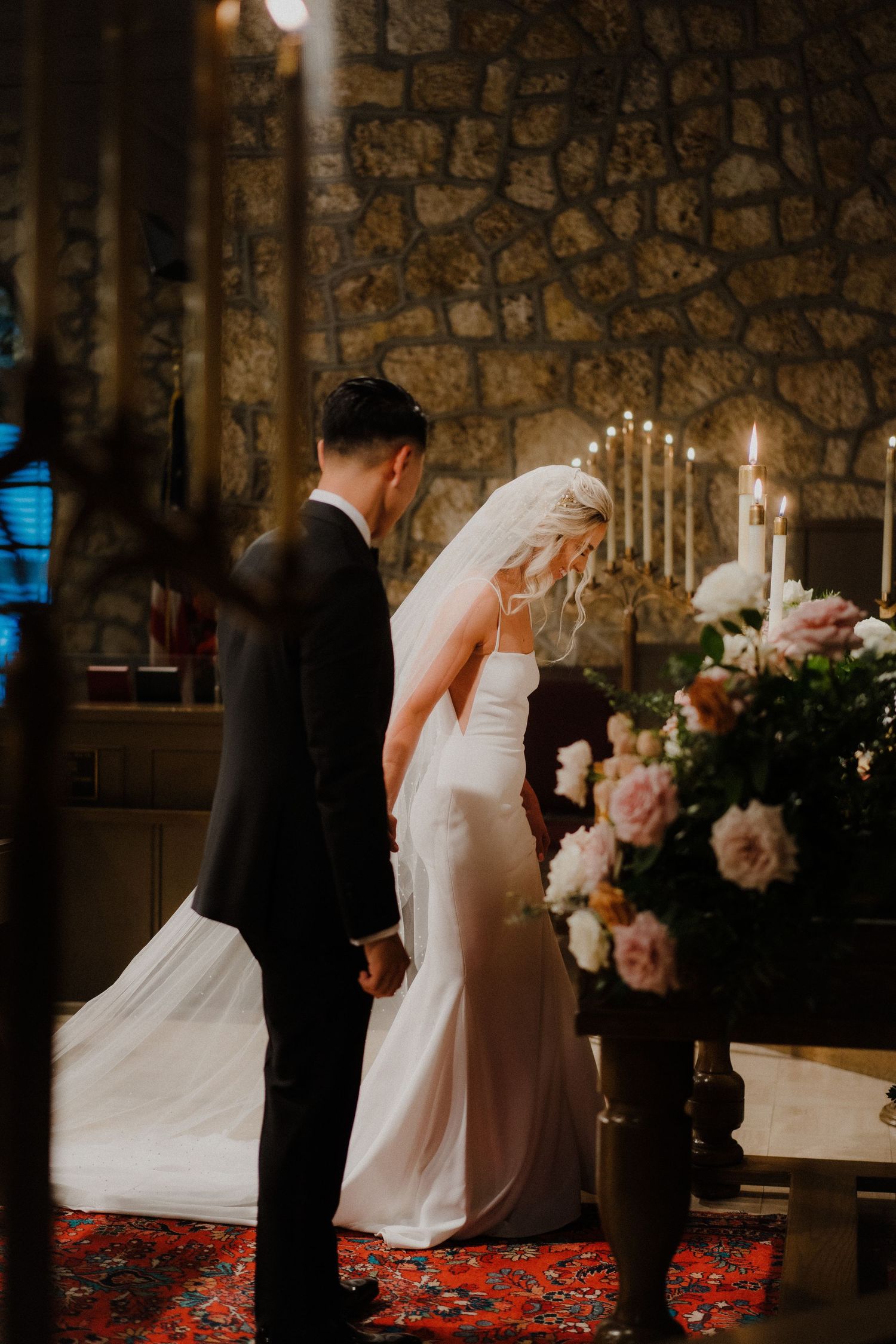 A bride and groom stand at the altar of a candlelit stone church, surrounded by lush floral arrangements during their wedding ceremony.