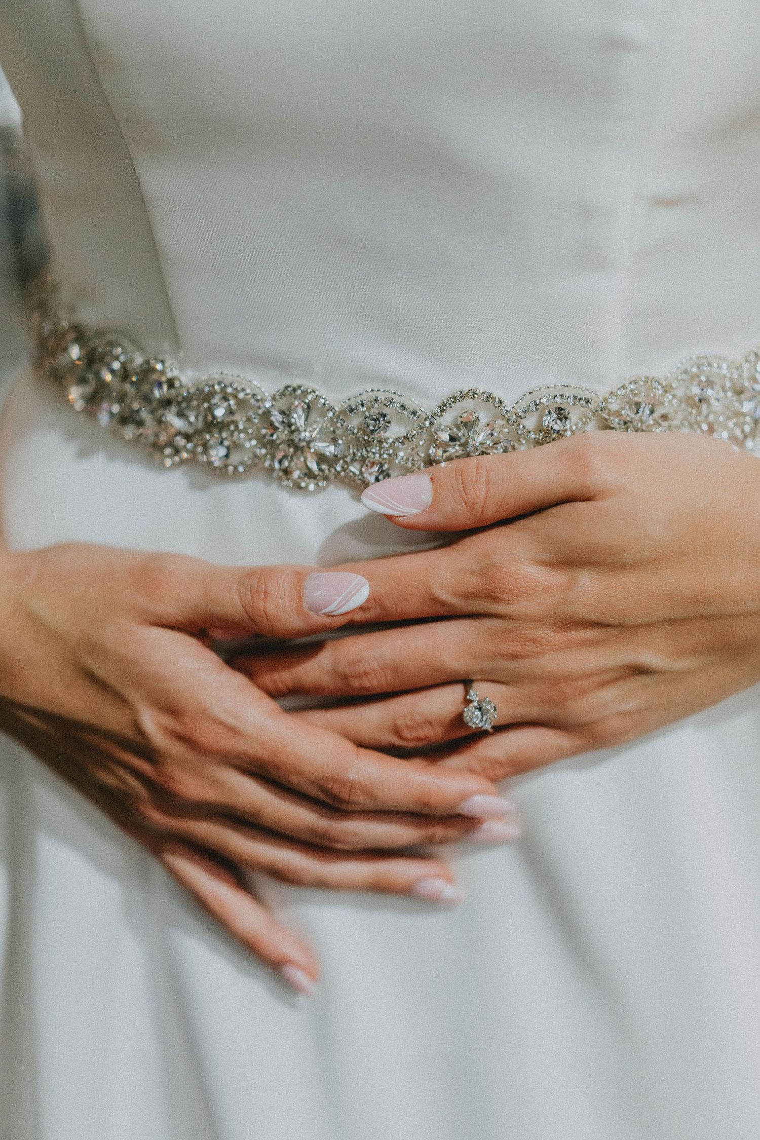 Close-up of bride's hands with elegant diamond engagement ring and French manicure on white beaded wedding gown.