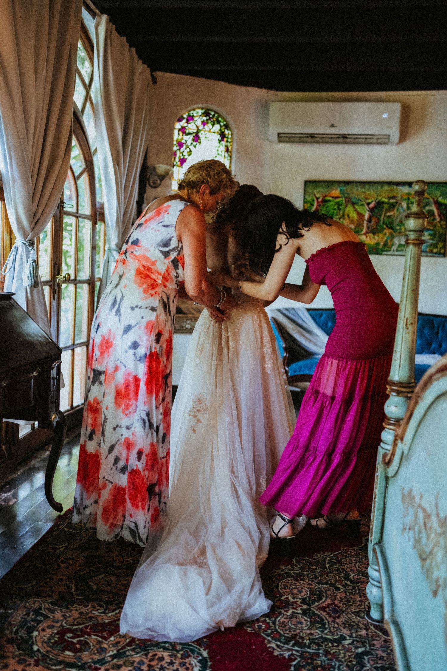 Two bridesmaids helping a bride into her wedding dress in an ornate room with stained glass windows.