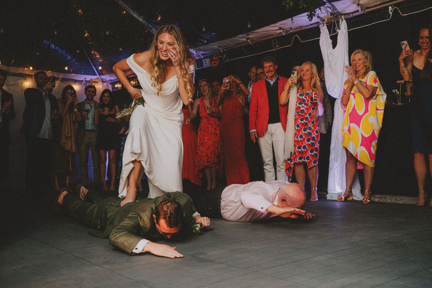 Bride in white dress stands over guests doing the worm on the wedding reception dance floor.