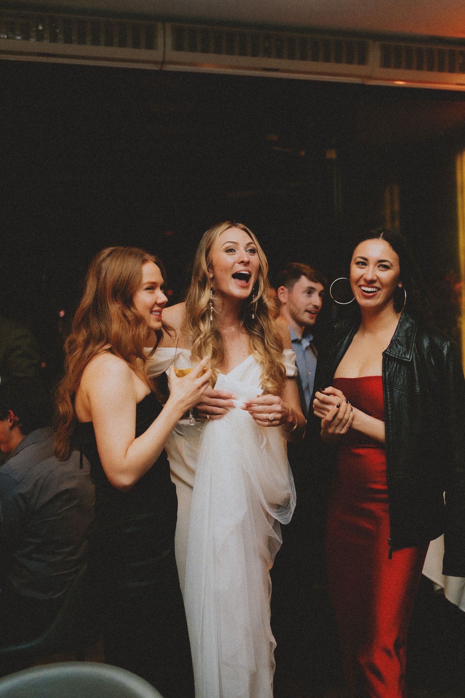 Bride in white slip dress laughs joyfully with two friends in black and red at wedding reception.