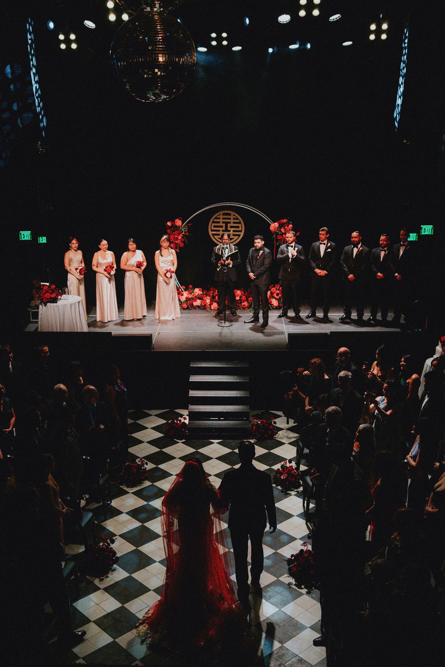 Wedding ceremony on a dramatic stage with bridesmaids, groomsmen, and couple on checkered floor with red floral decor.