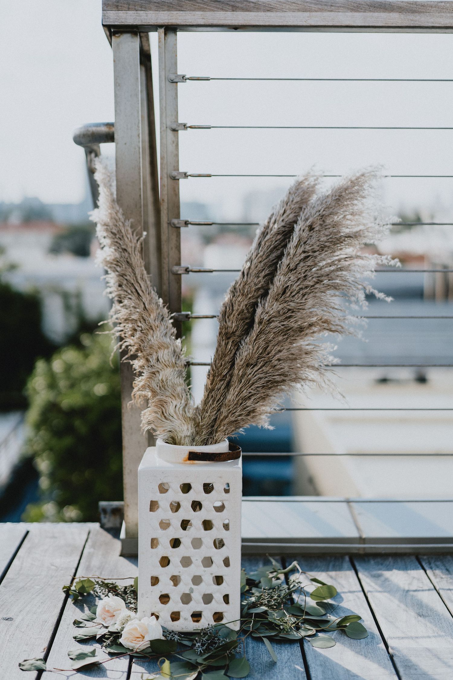 White lattice lantern vase with pampas grass on wooden deck, surrounded by greenery and white roses.