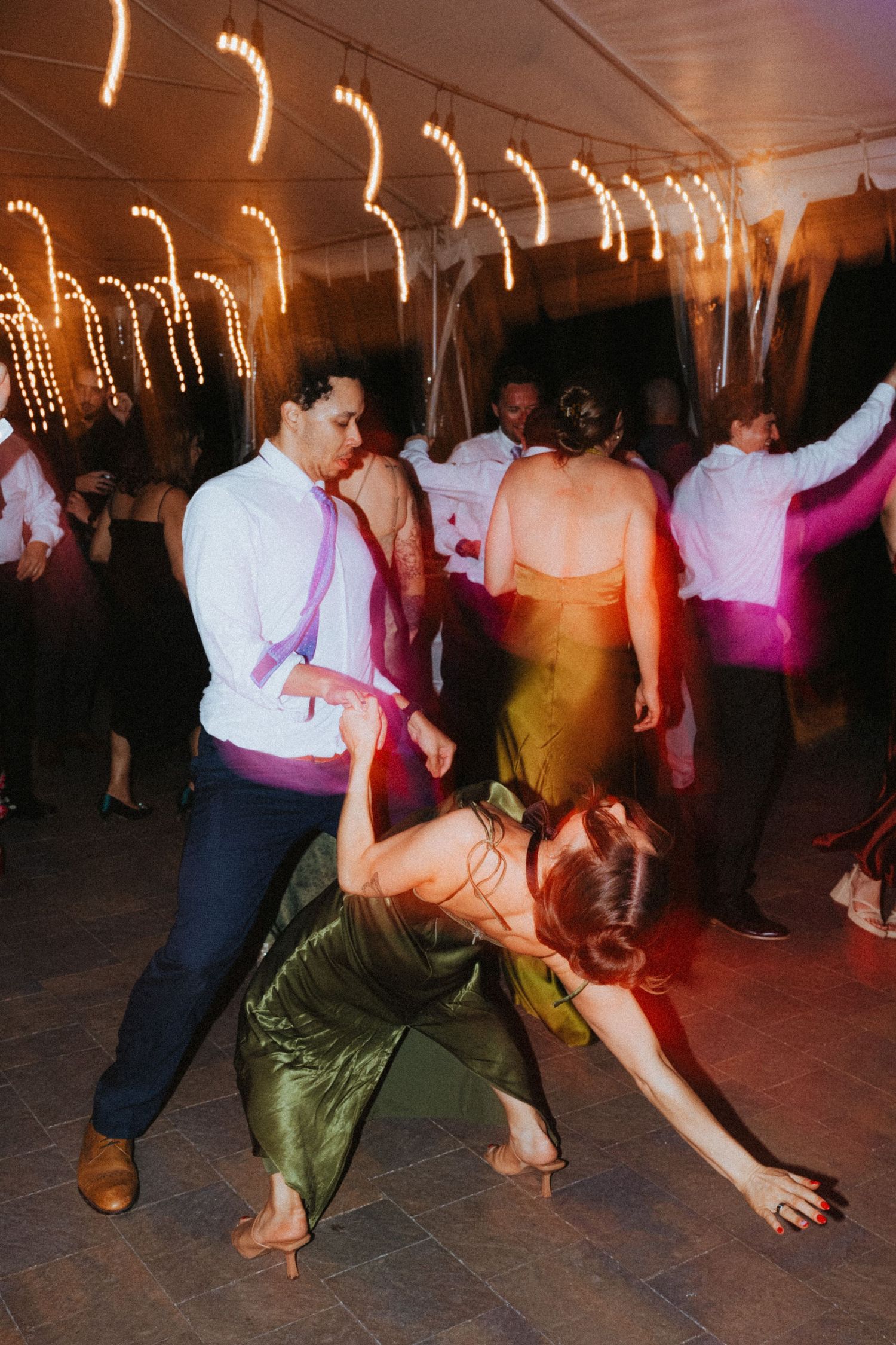 Wedding guests dance energetically under string lights at a lively outdoor wedding reception.
