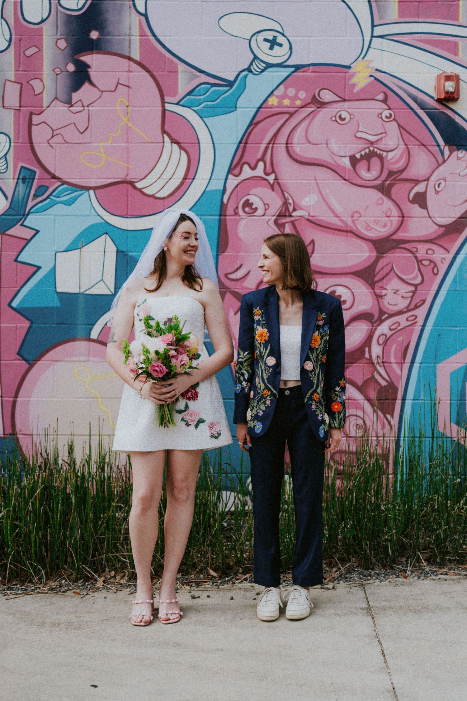 Two brides smile together in front of a vibrant pink street mural, one holding a colorful bouquet.
