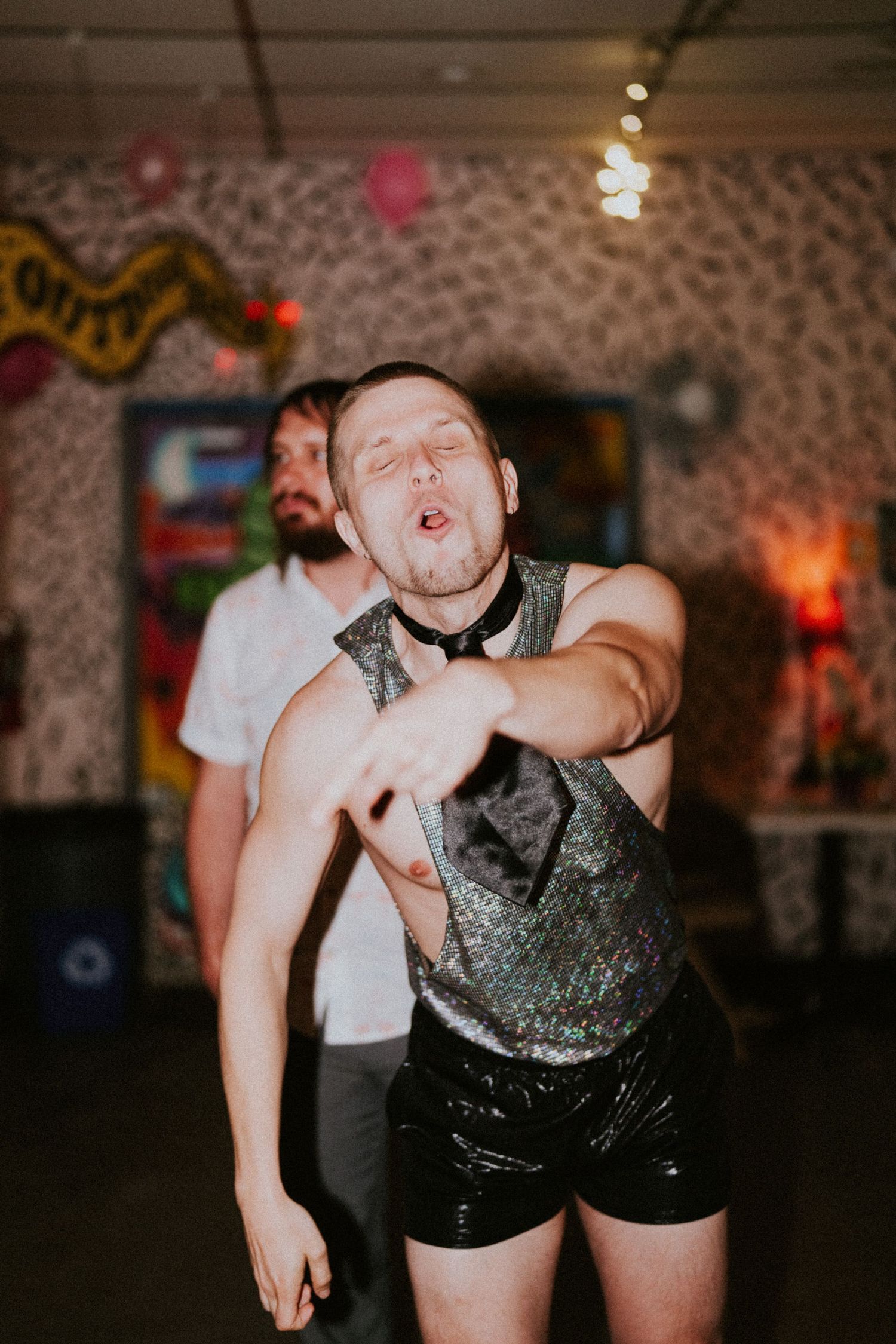 Man in sequin vest and black shorts striking a bold dance pose at an indoor party event.