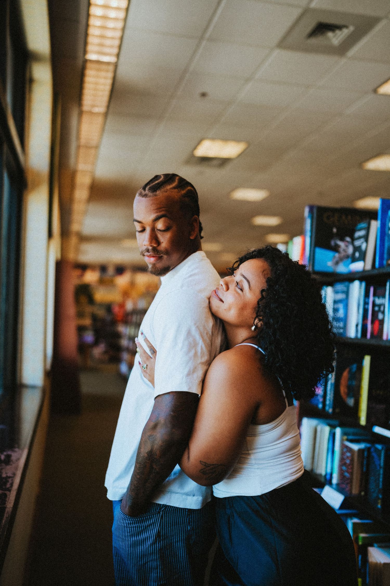 A couple embraces romantically in a bookstore, woman resting her head on mans back.