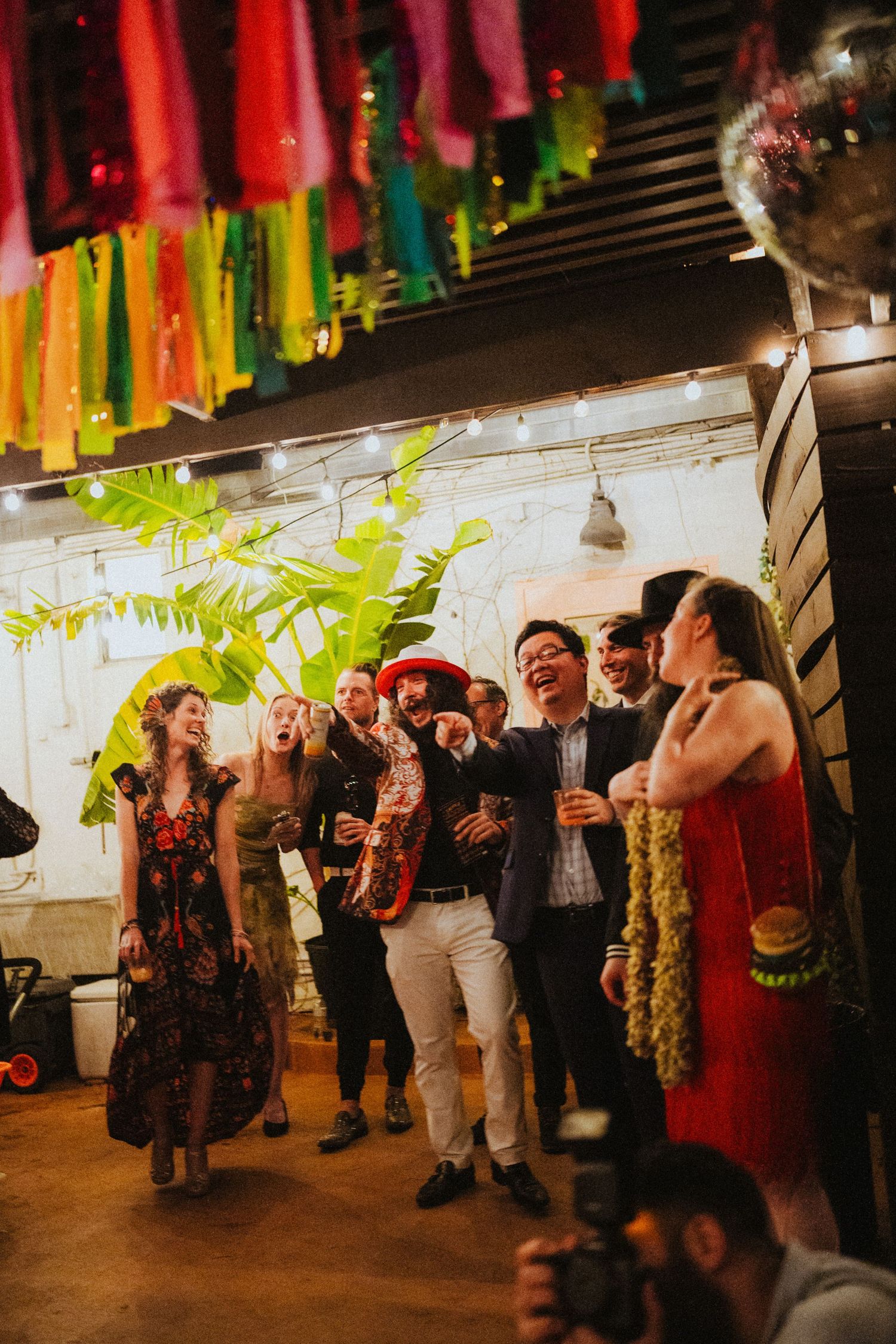 Colorful fiesta wedding reception with guests laughing under vibrant paper fringe decorations and string lights.