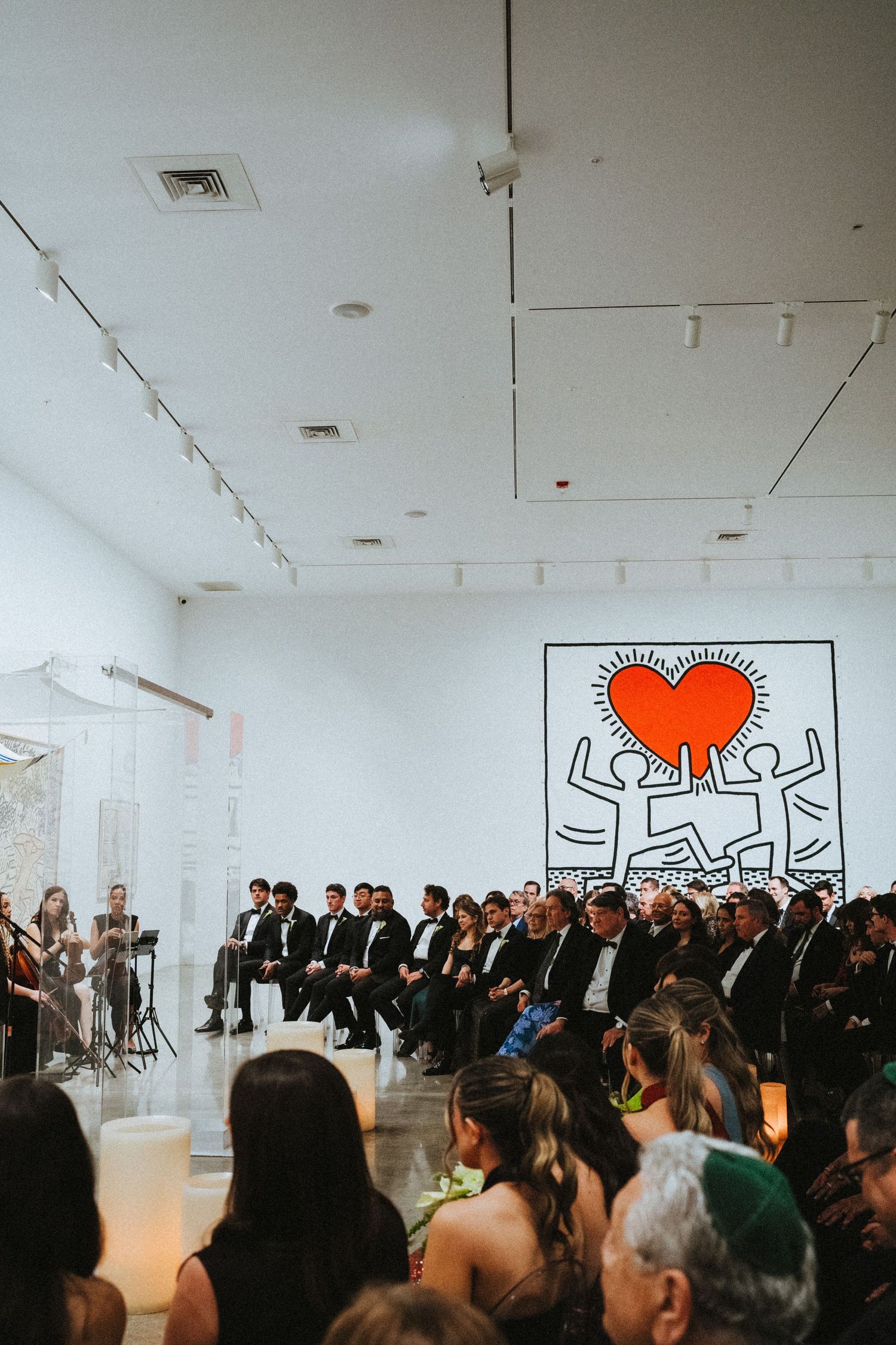 Jewish wedding ceremony in art gallery with Keith Haring heart mural, guests in formal attire seated in rows.