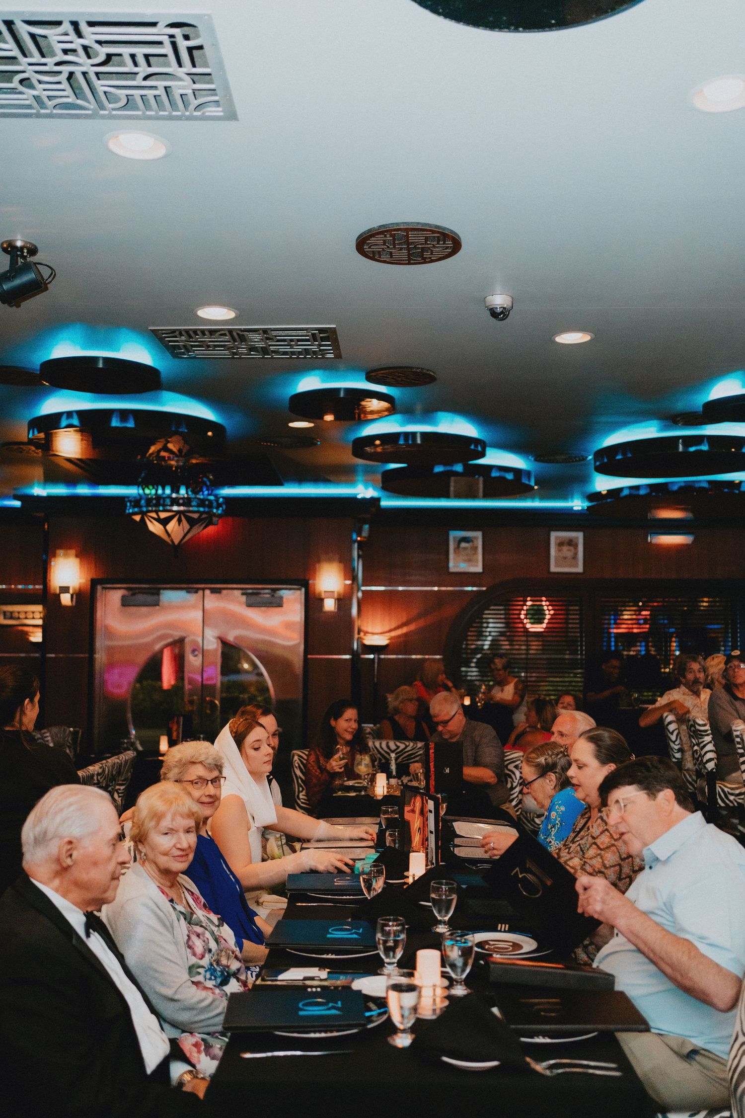 Guests dining at an elegant restaurant with blue ambient lighting and decorative ceiling fixtures during a wedding reception.